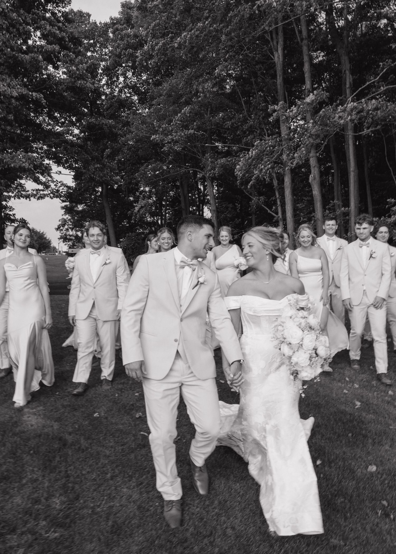 Wedding party walking and laughing together across green lawn in all-white outfits.