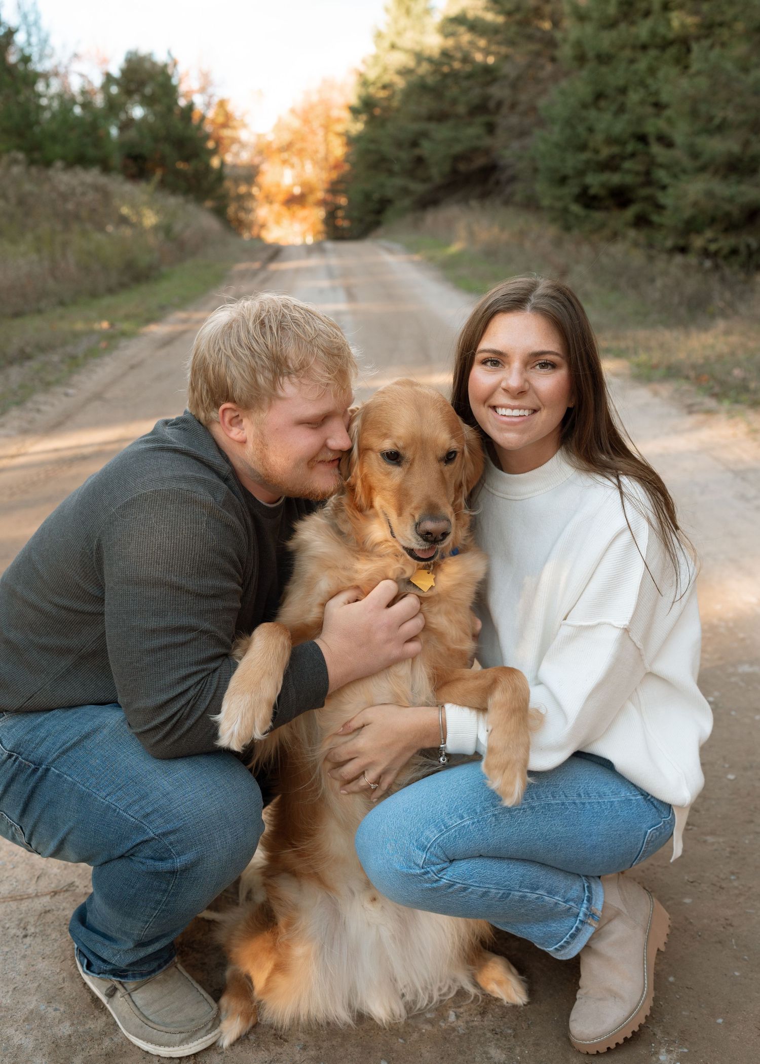 A couple and their golden retriever share a loving moment on a dirt road at sunset.