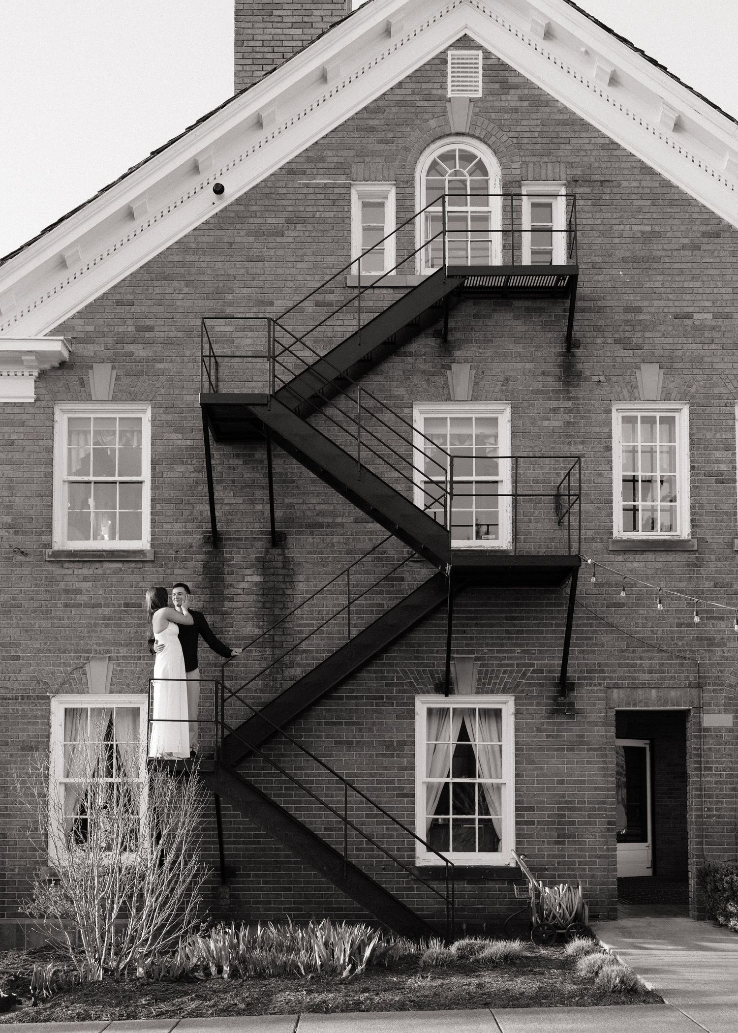 Couple poses on brick building fire escape staircase in black and white wedding portrait.