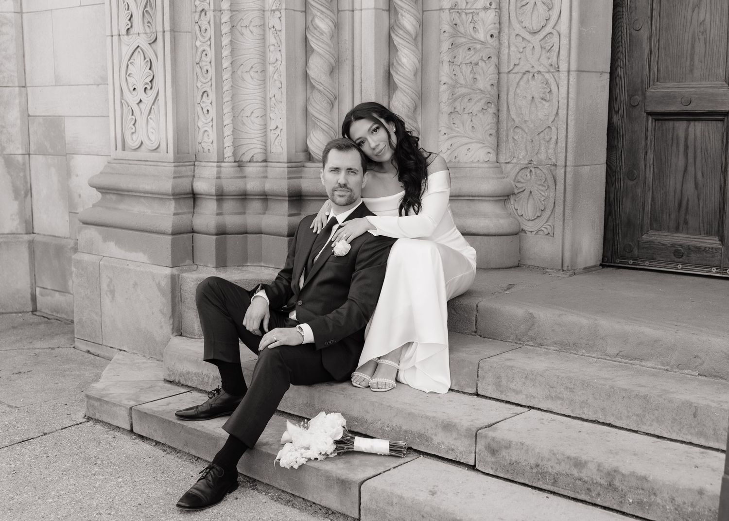 Black and white photo of a bride and groom sitting on church steps in elegant wedding attire.