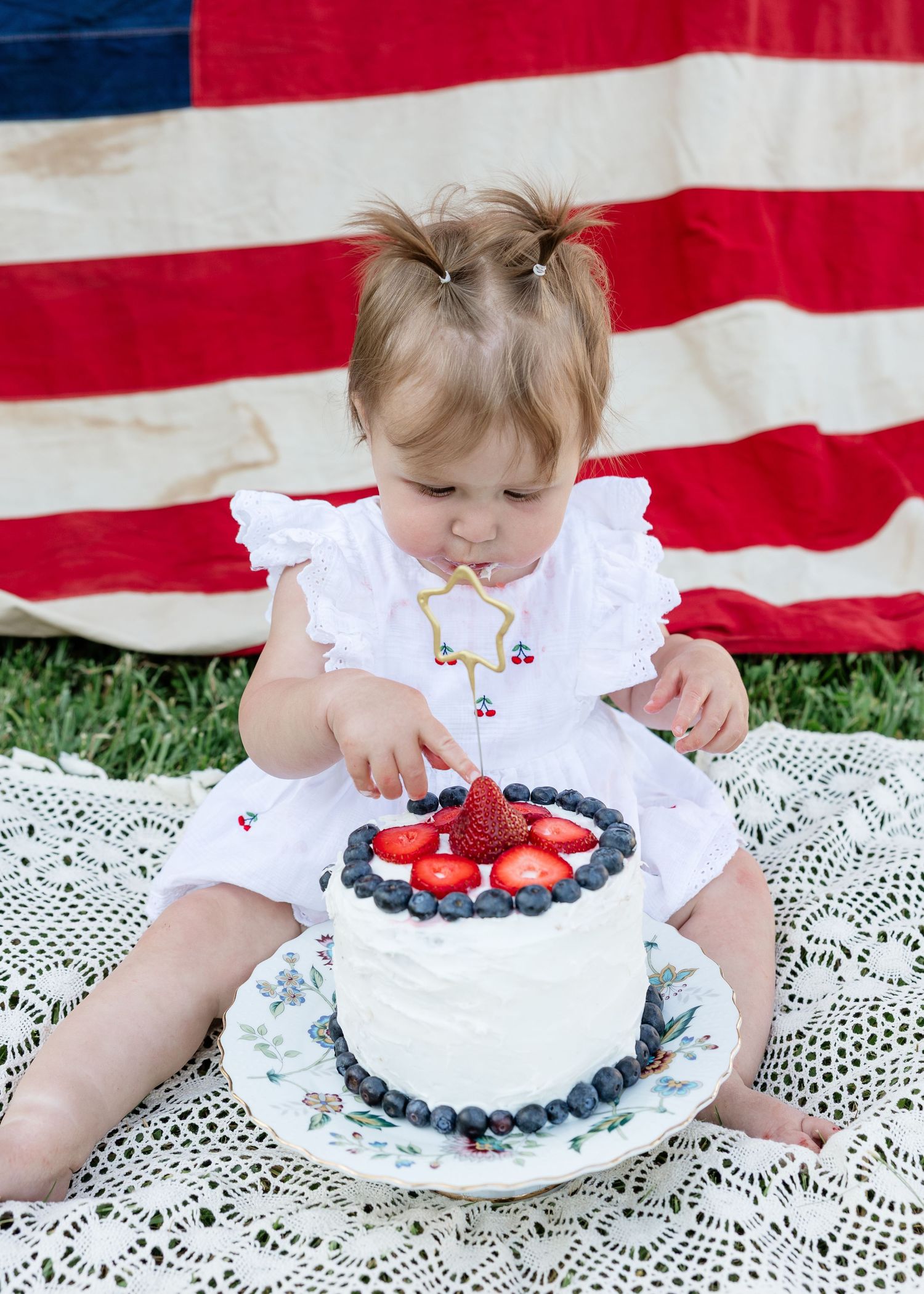 Baby girl in white dress touching berry-topped birthday cake outdoors with American flag backdrop.