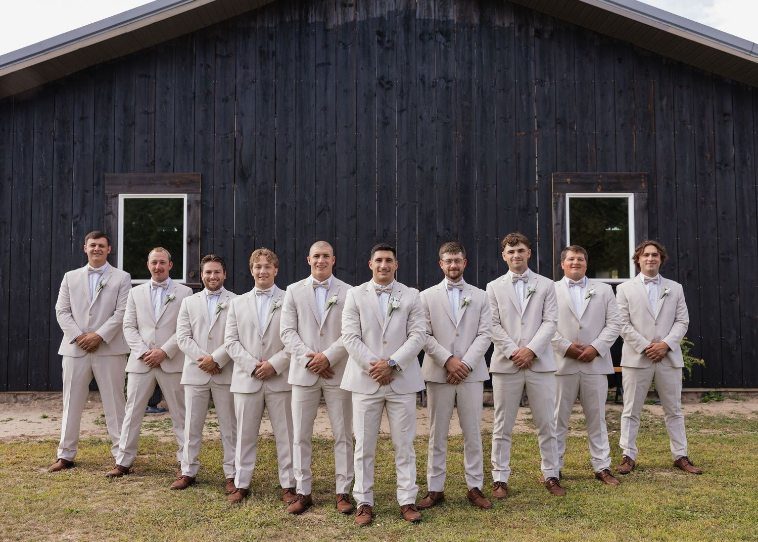 Wedding party in white attire poses in front of black barn building.