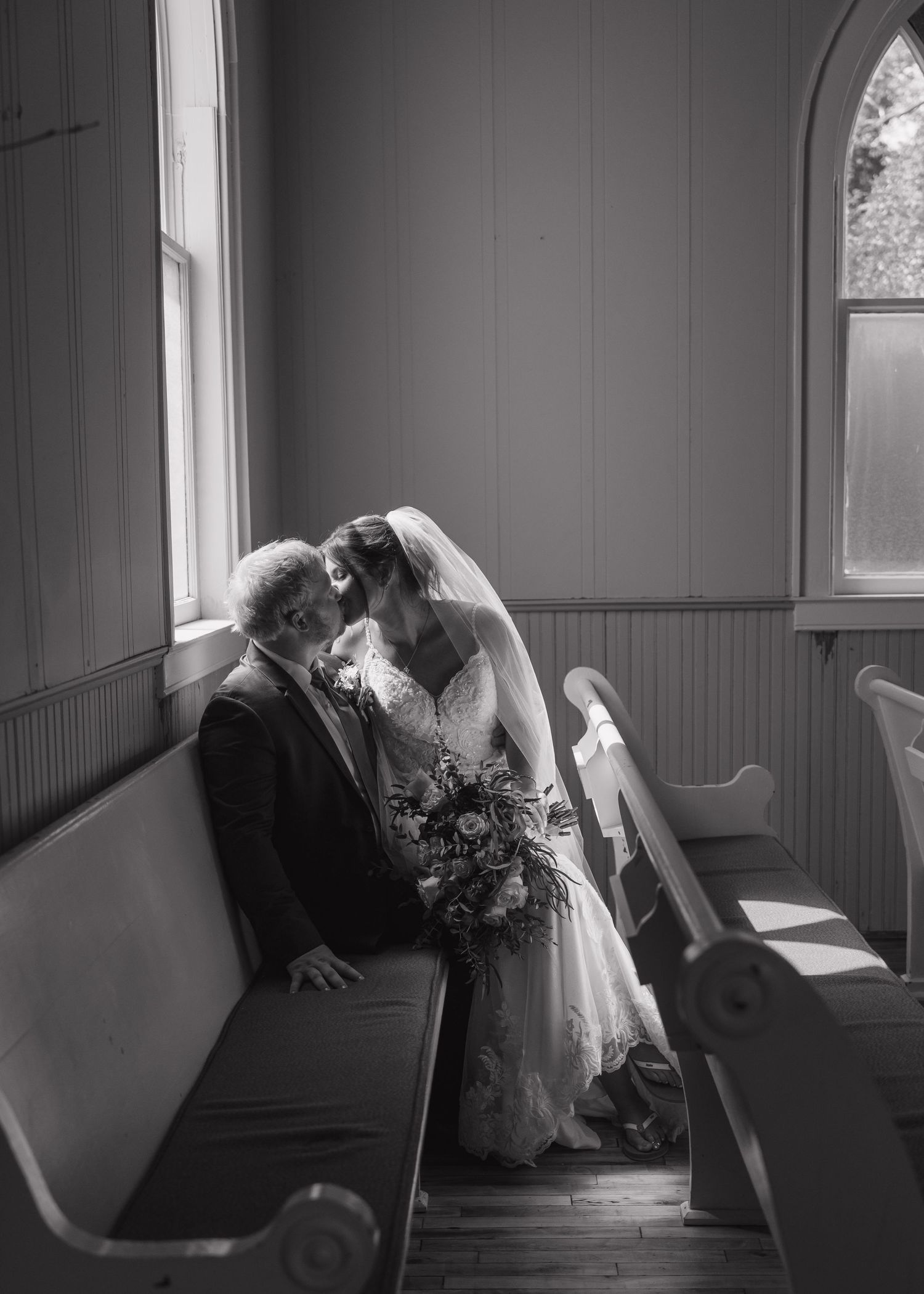 A romantic black and white moment between newlyweds in a church pew by a window.