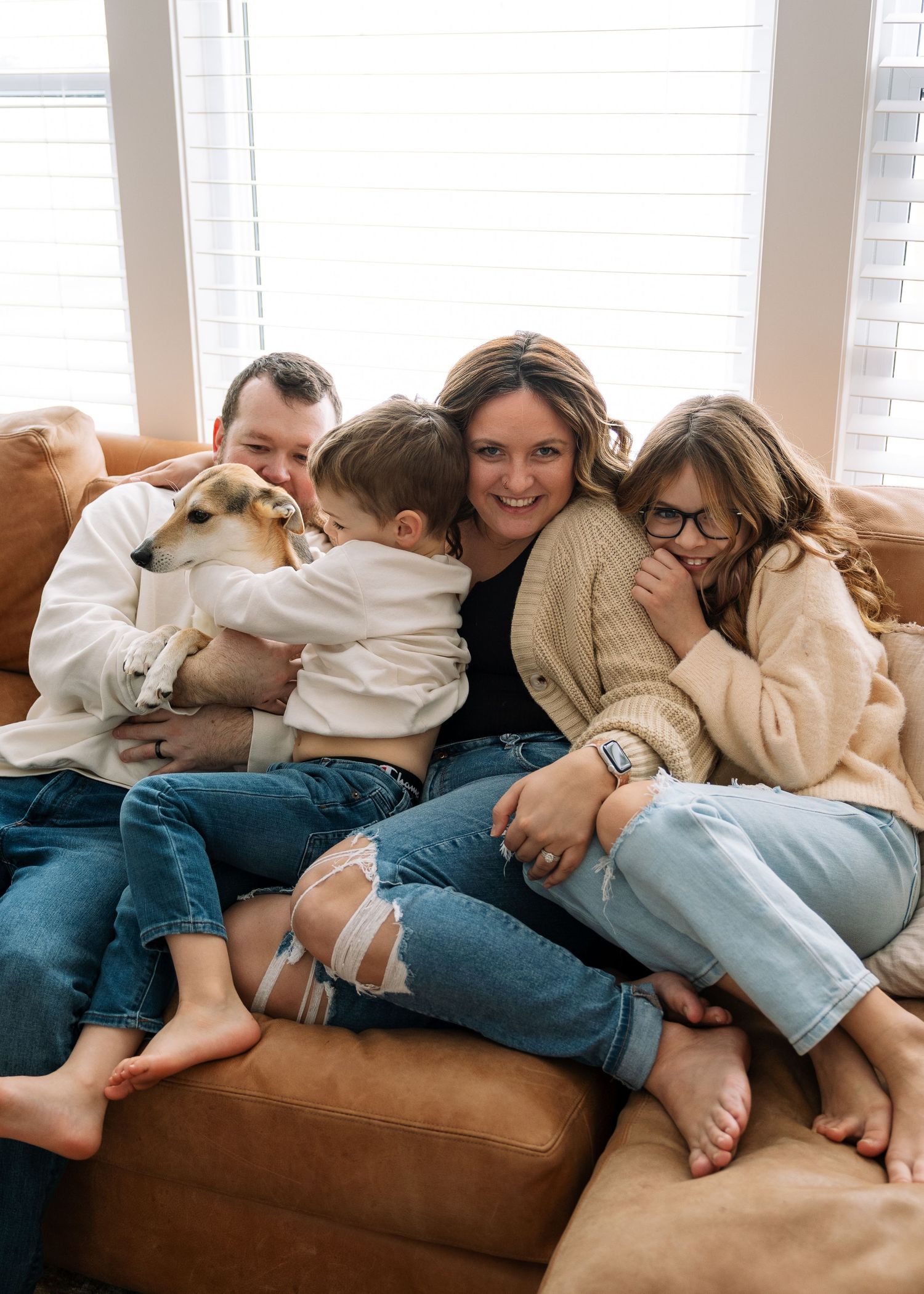 A family cuddles together on a brown leather couch with their pet dog.