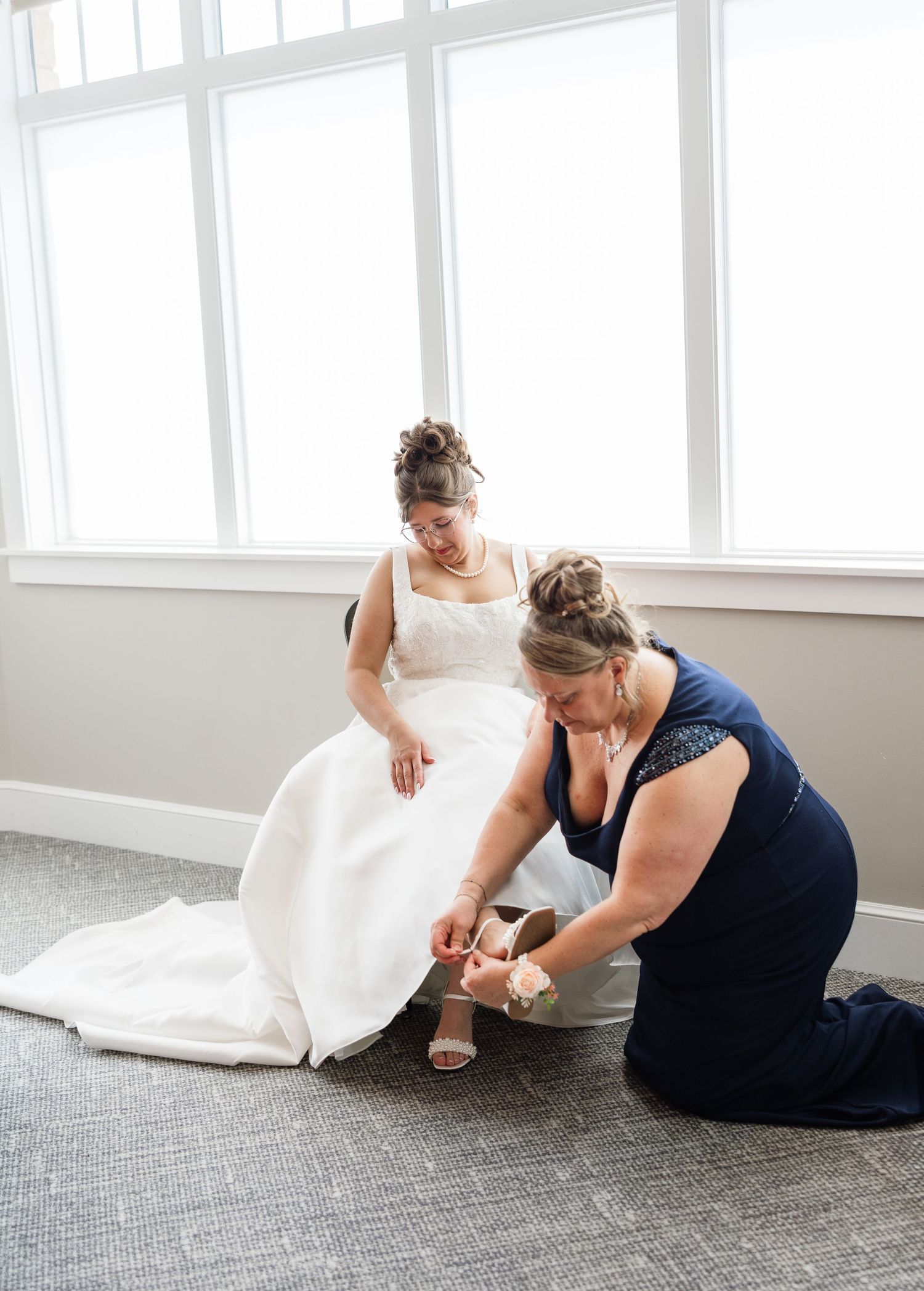 A bridesmaid in navy blue kneels to help a bride in a white gown put on her shoes before the wedding ceremony.