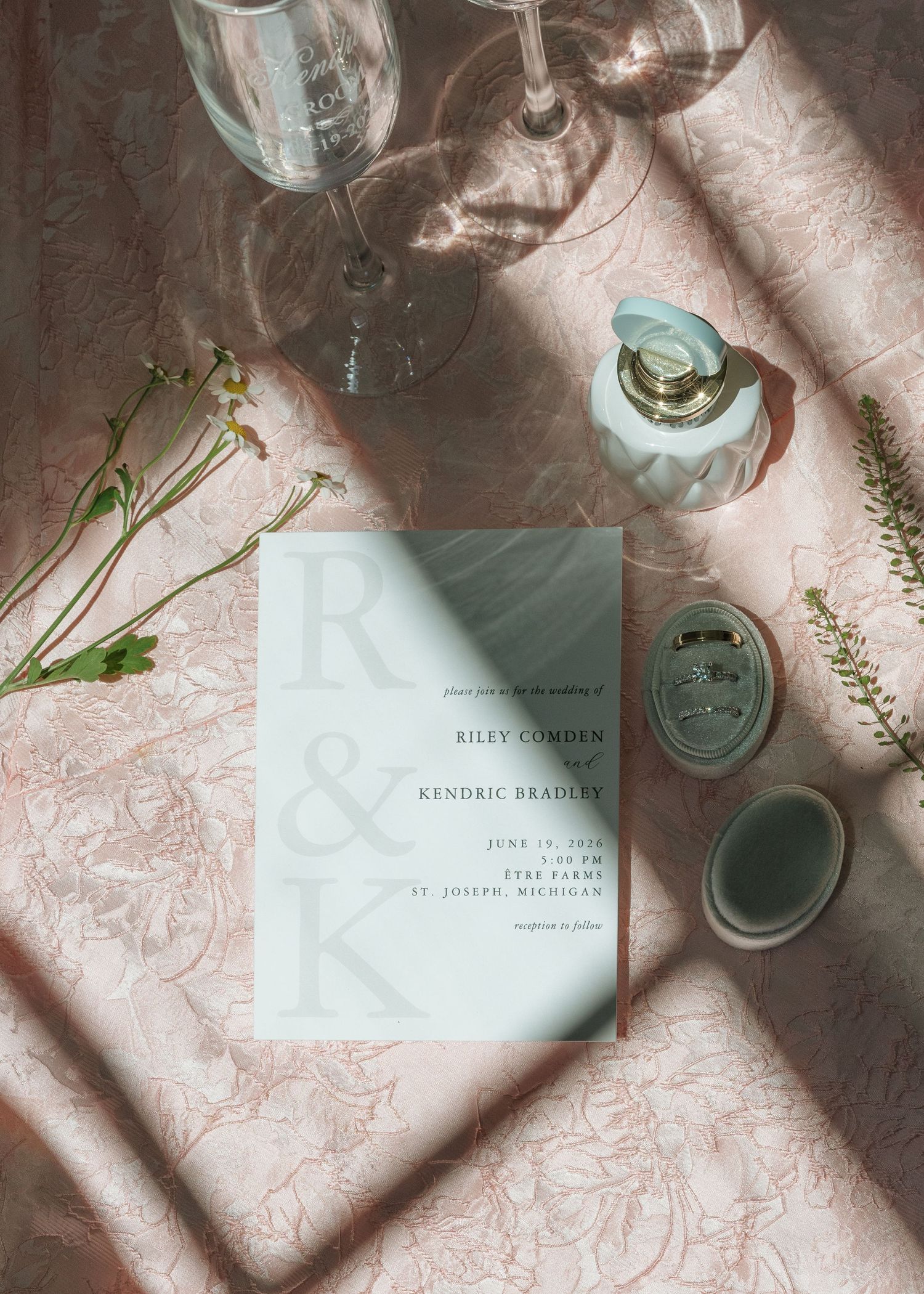 Elegant wedding invitation flat lay with monogram initials, wine glasses, ring box, and floral accents on pink linen.
