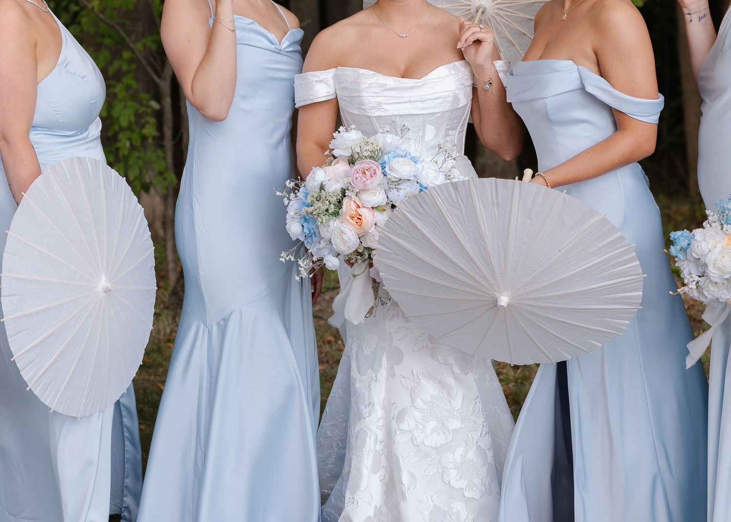 Bridesmaids in light blue dresses hold white parasols during outdoor wedding photos.