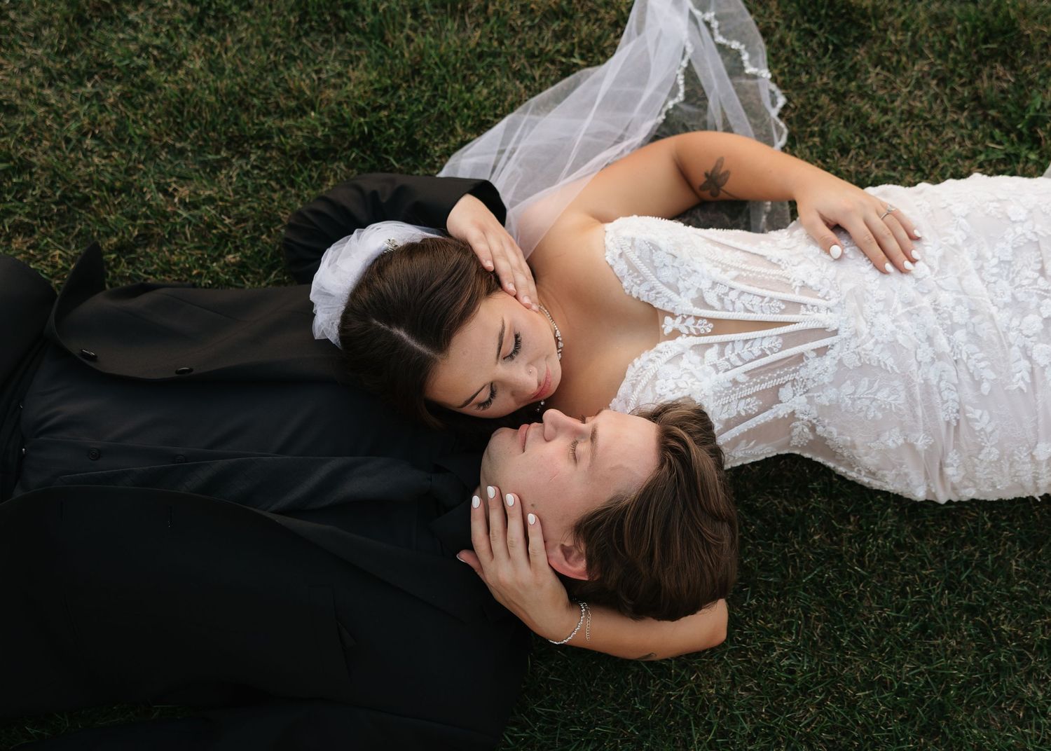 A tender moment between newlyweds lying on grass during their wedding photoshoot.