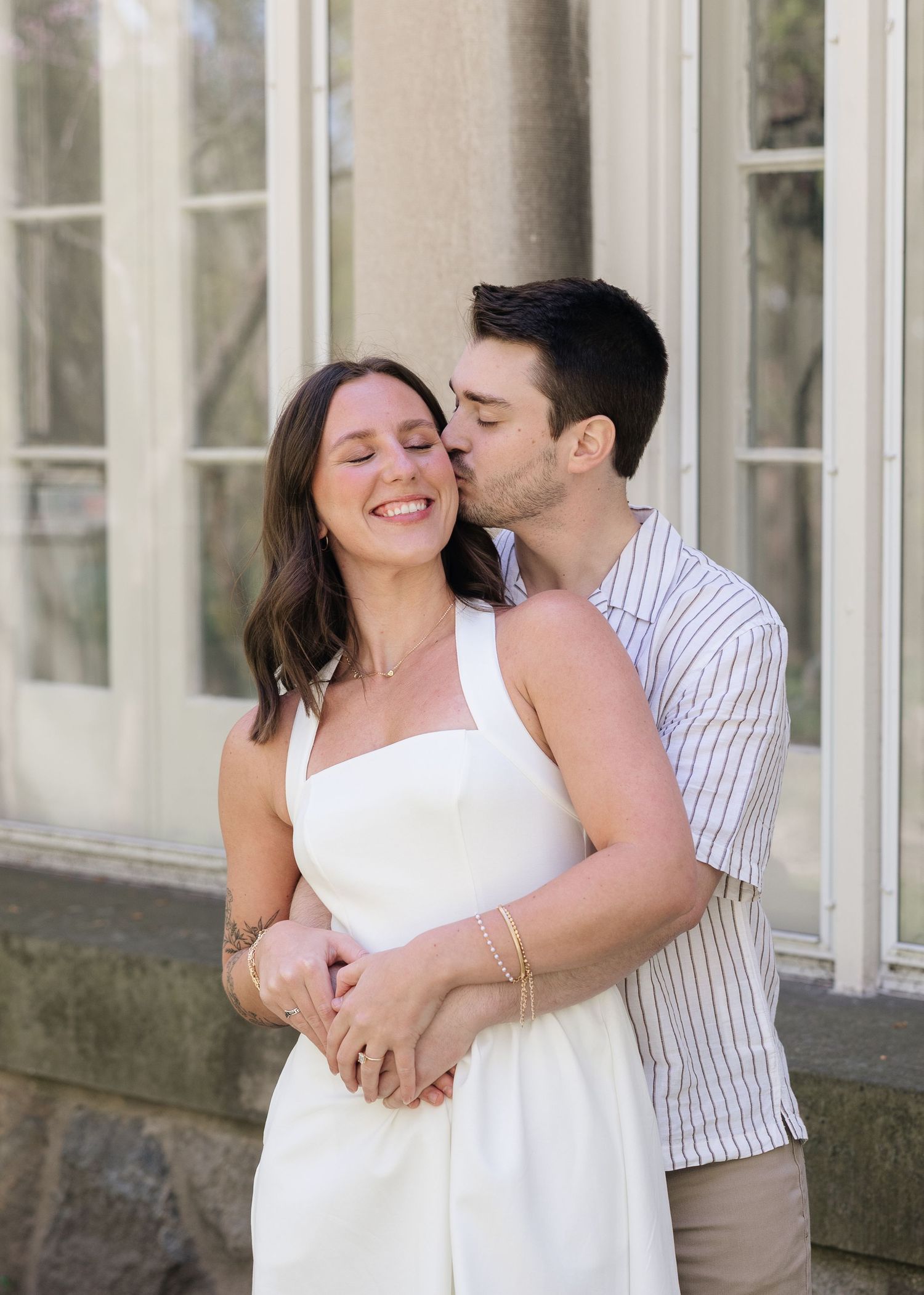A couple shares a tender moment outdoors, man in striped shirt kissing womans cheek as she smiles in white dress.
