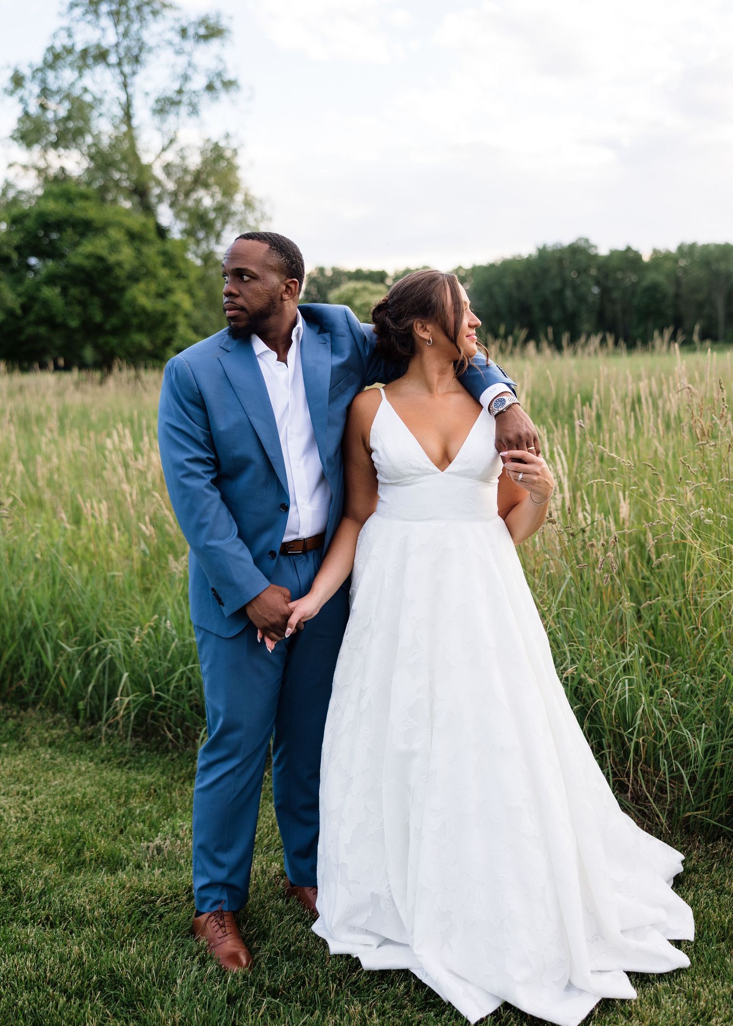 Groom in blue suit and bride in white gown pose together in lush green field on wedding day.