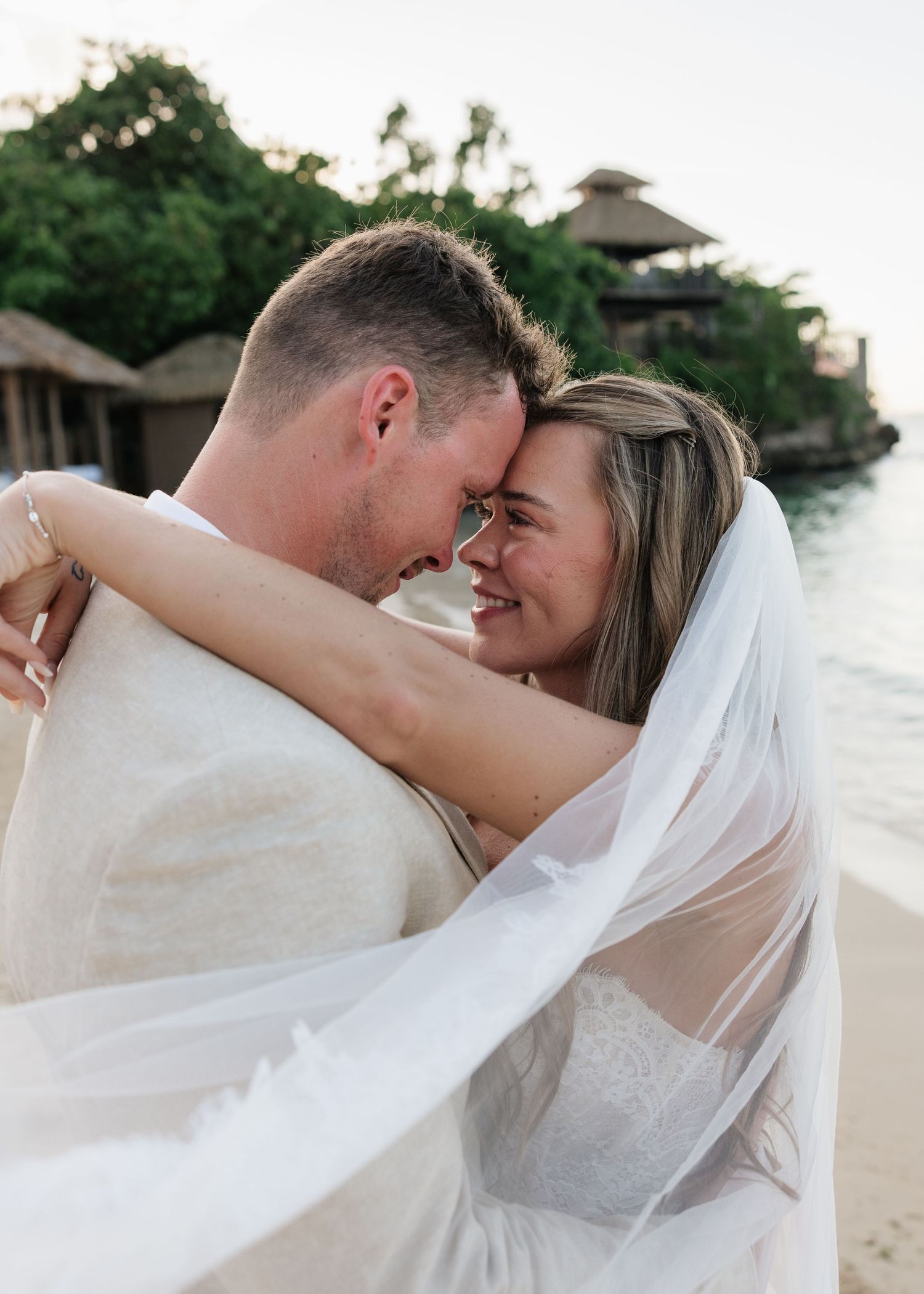 A couple in wedding attire embraces on a beach, the bride in a flowing veil smiling at her groom near tropical waters.