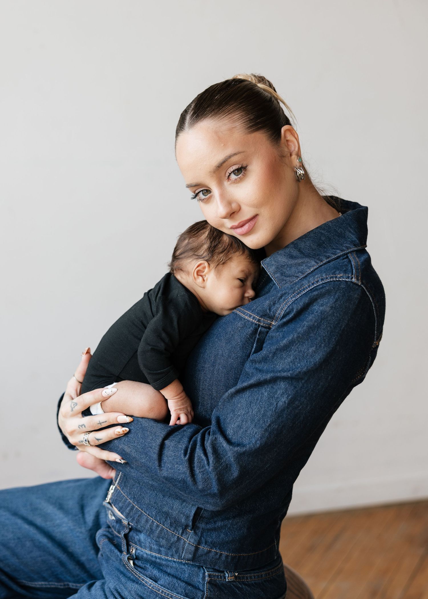 Young woman in denim jumpsuit tenderly holding a sleeping newborn baby against her chest in a studio setting.