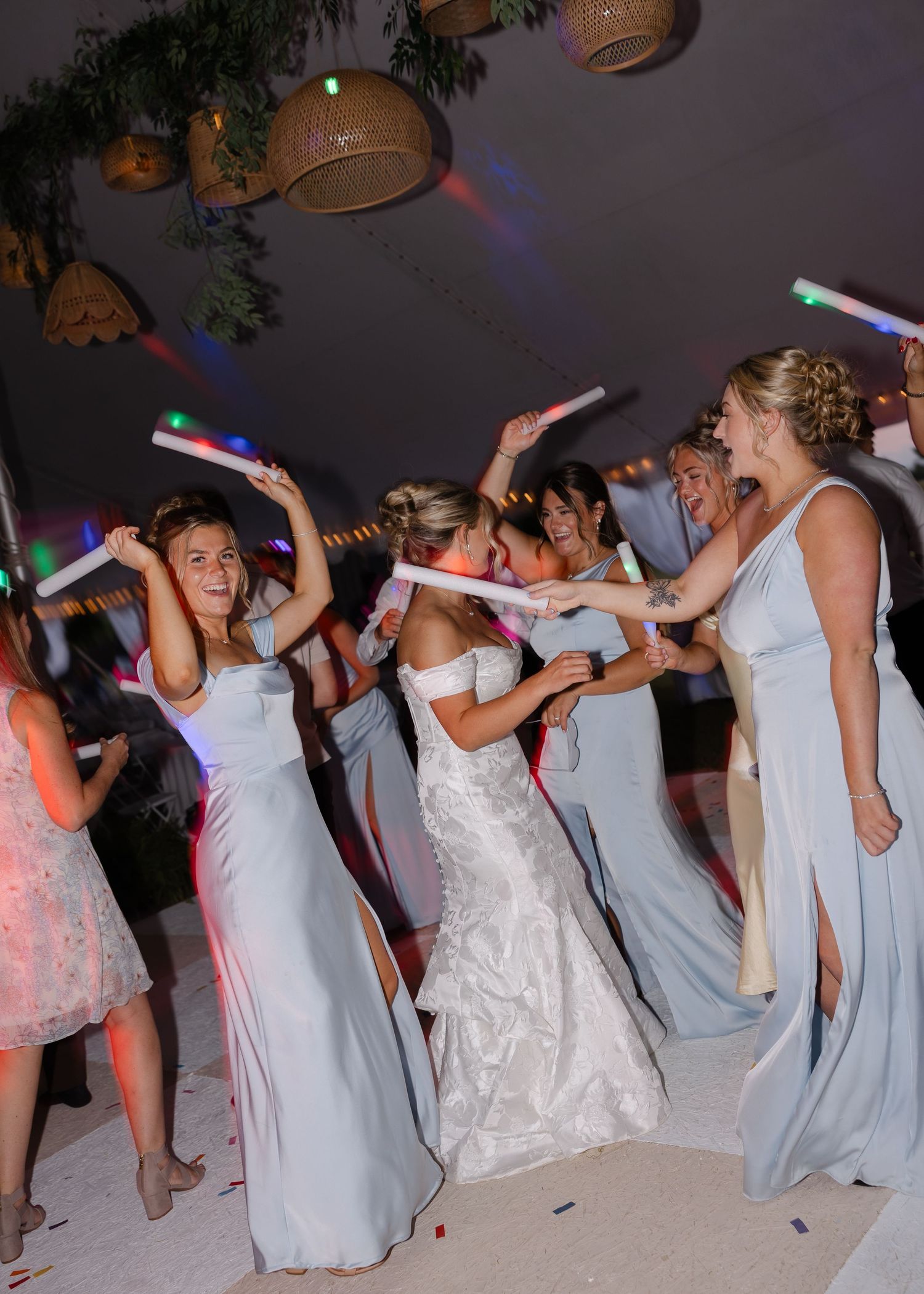 Wedding party dances in a line holding glow sticks under a tent at night.