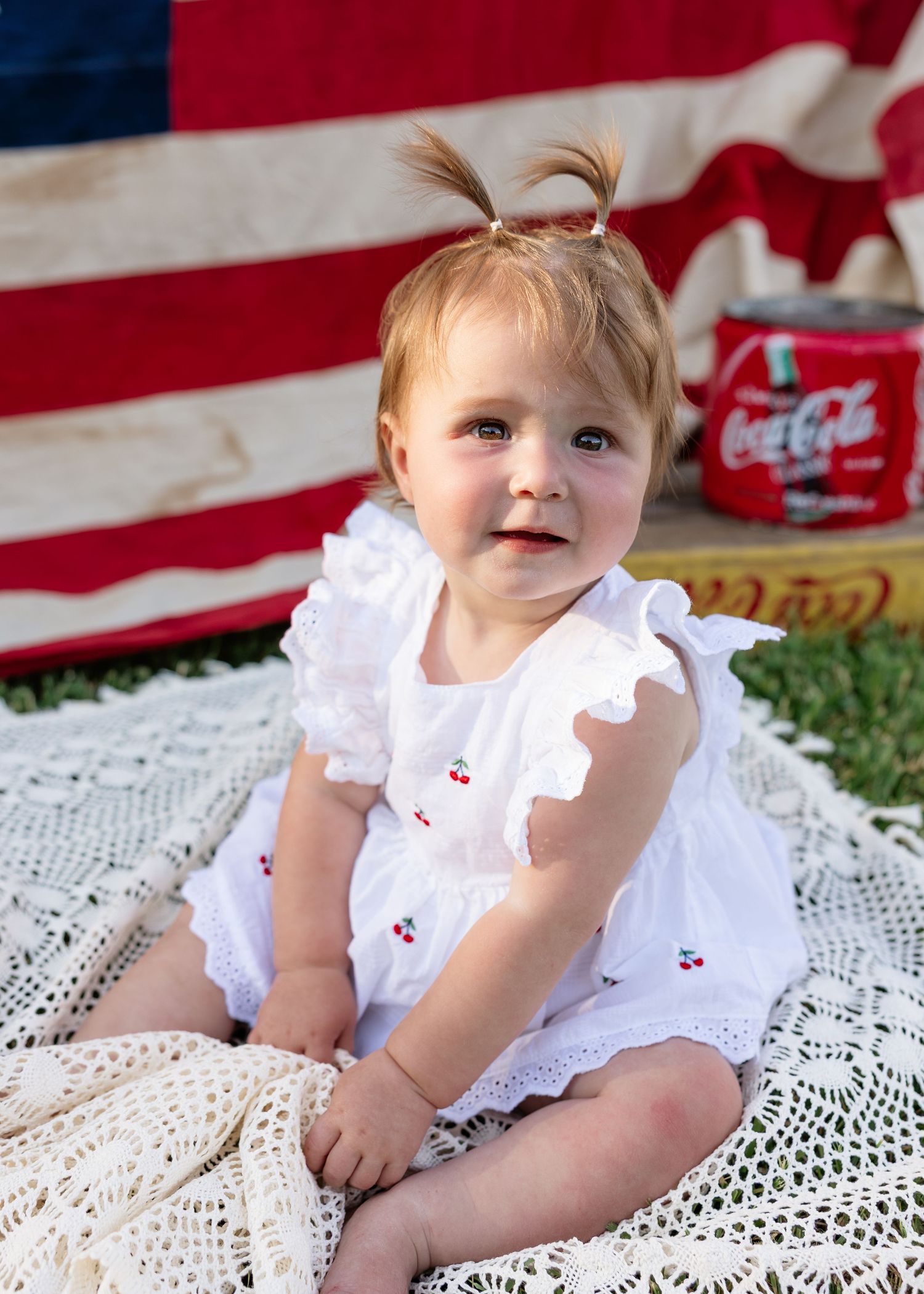 Adorable baby girl in white cherry dress sits on lace blanket with American flag and Coca-Cola can backdrop.