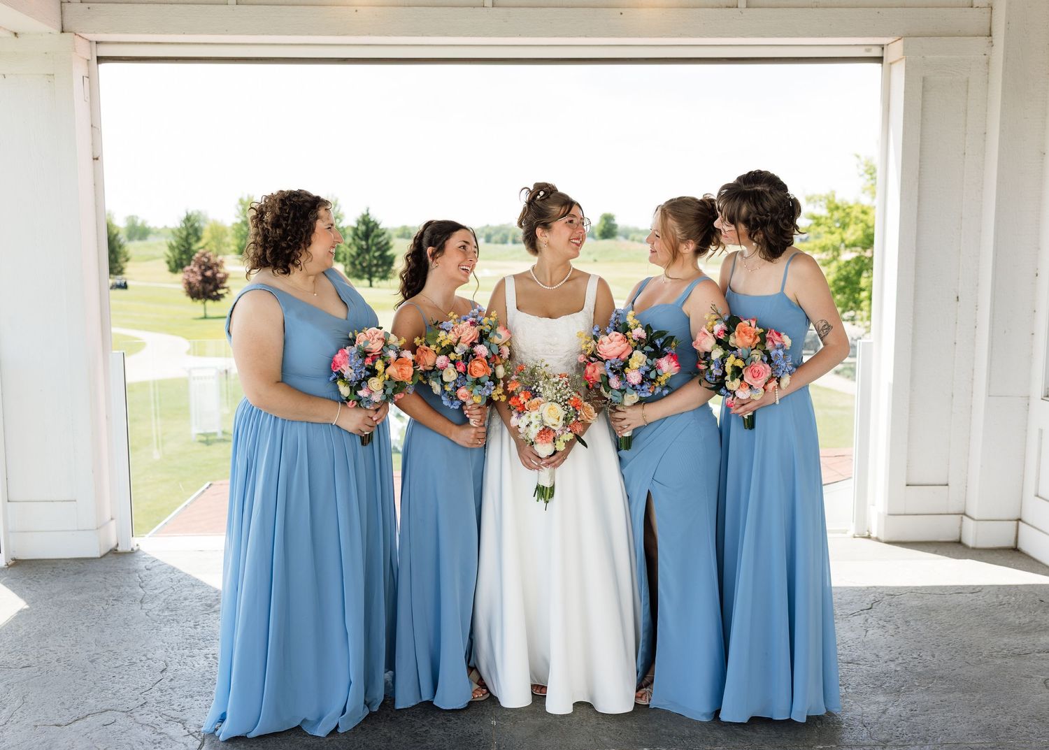 Bride in white gown with four bridesmaids in blue dresses holding colorful bouquets at a wedding venue.