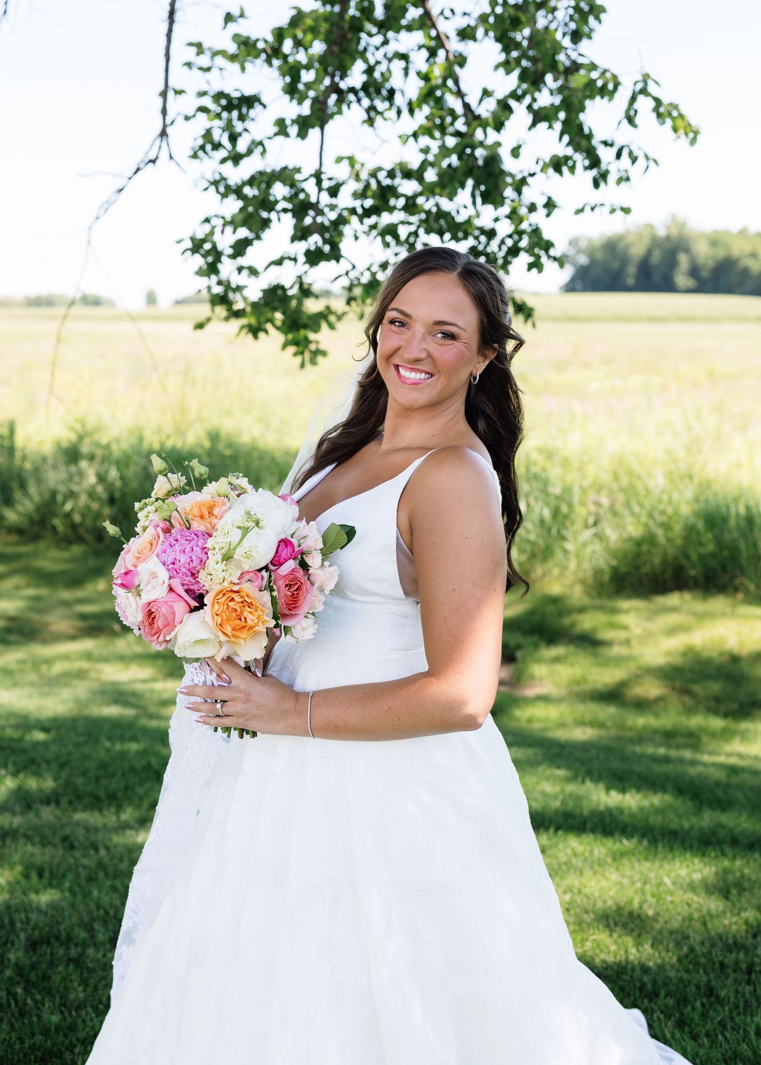 Smiling bride in white gown holding colorful pink and orange bouquet outdoors in green field on sunny wedding day.