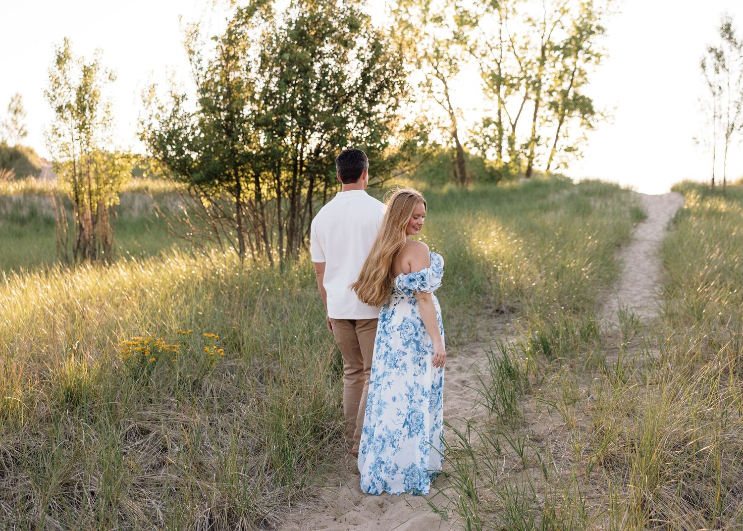 Couple walking hand in hand through a golden meadow path surrounded by tall grass and trees at sunset.