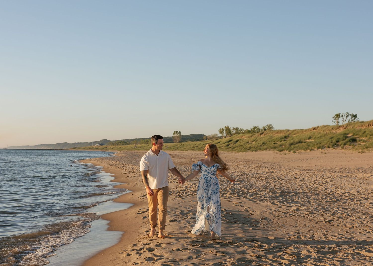 Couple holding hands walking along a sandy beach at golden hour with calm waves and green dunes in the background.