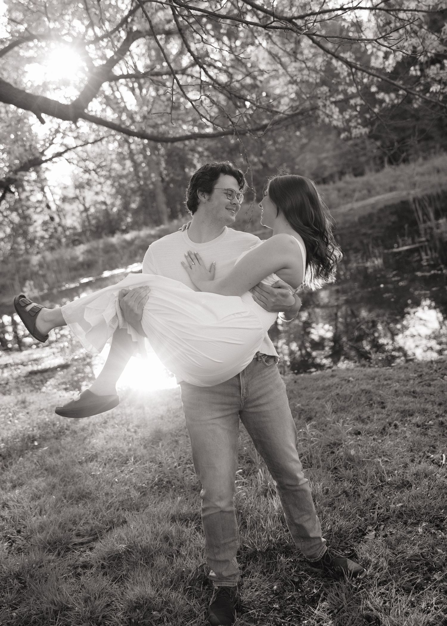 Black and white photo of a man carrying a woman in his arms outdoors near a pond, smiling at each other.