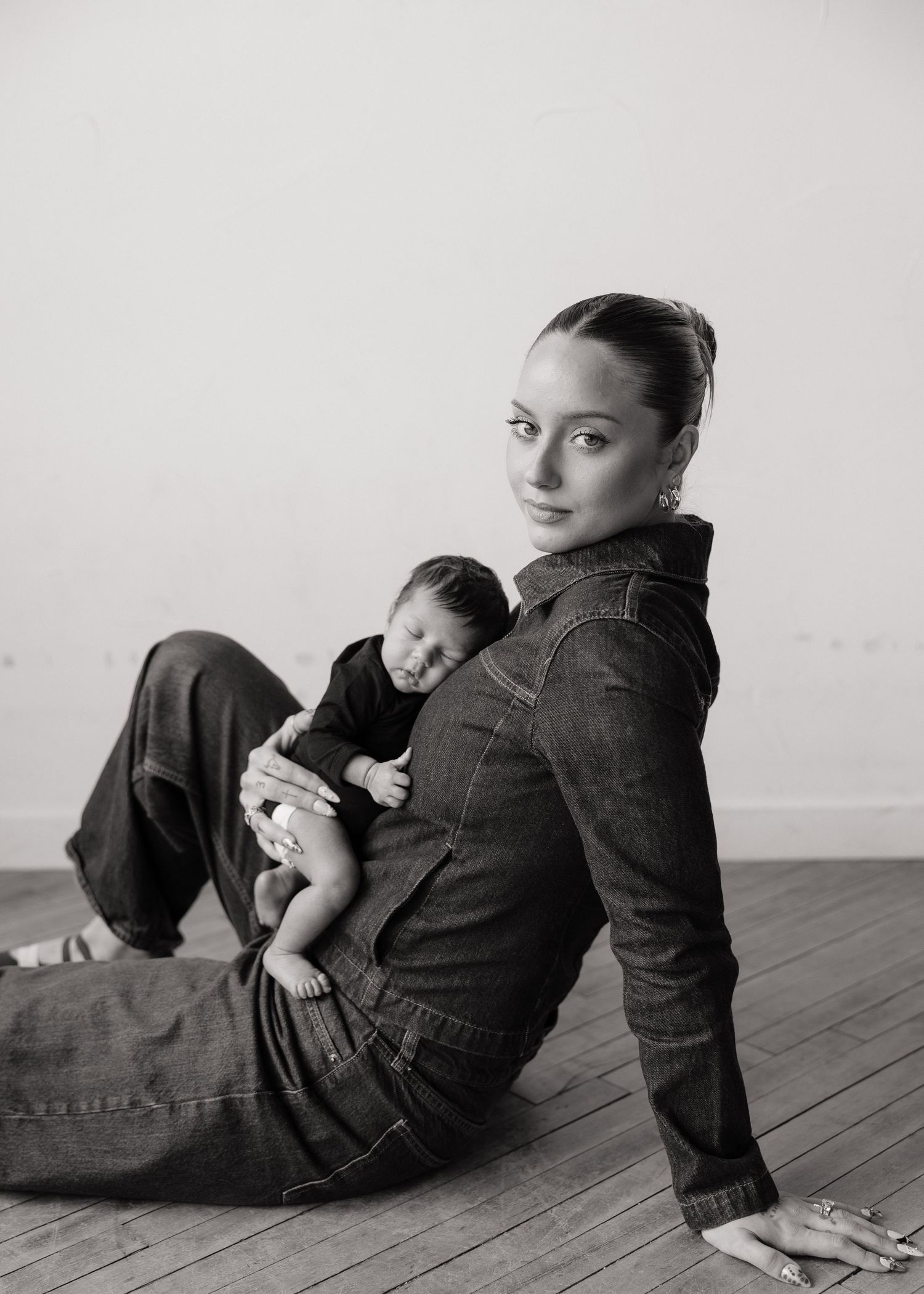 Black and white photo of a young woman in denim sitting on wood floor cradling a newborn baby.