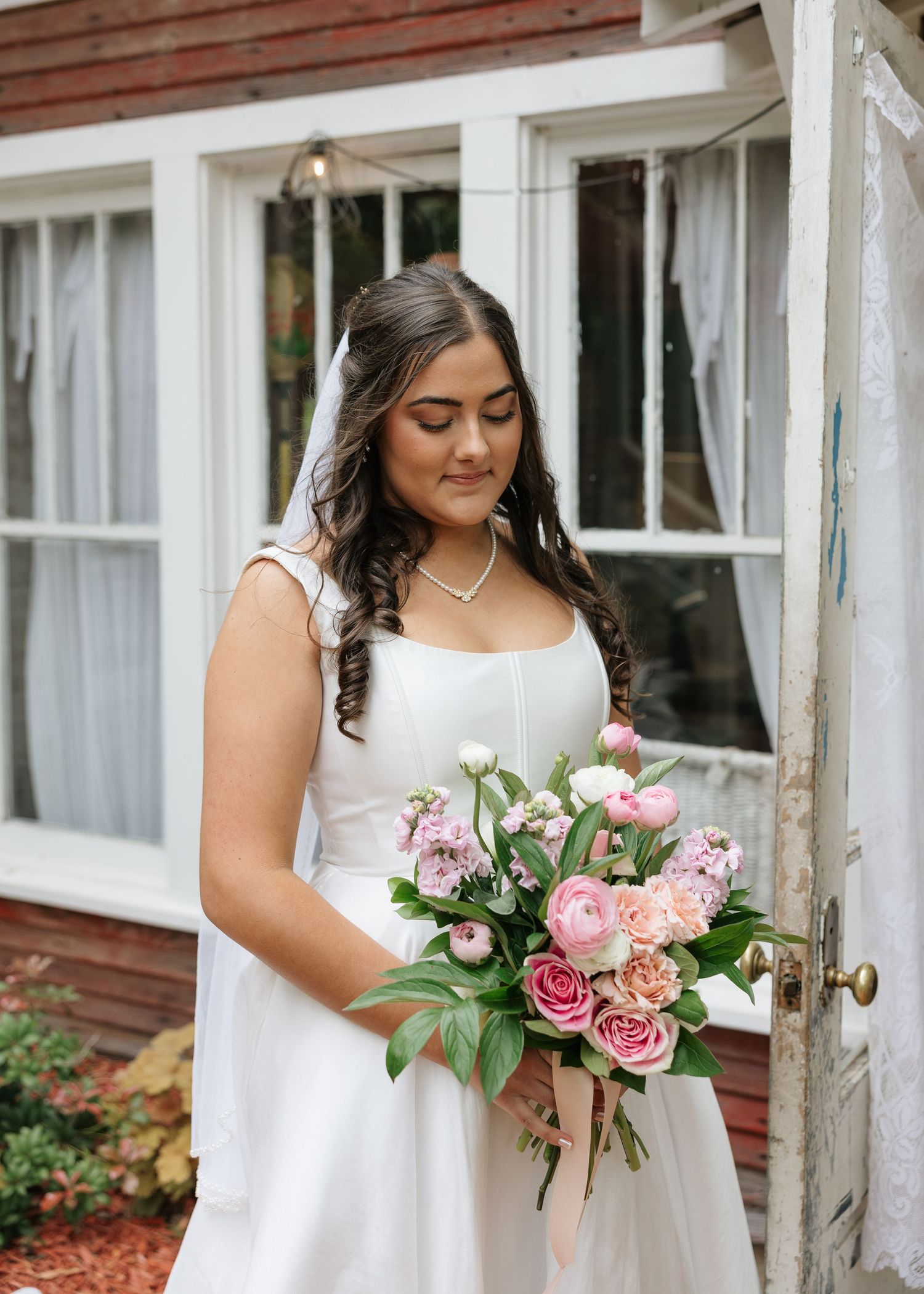 Bride in white gown holding pink floral bouquet stands before rustic red building with white windows.