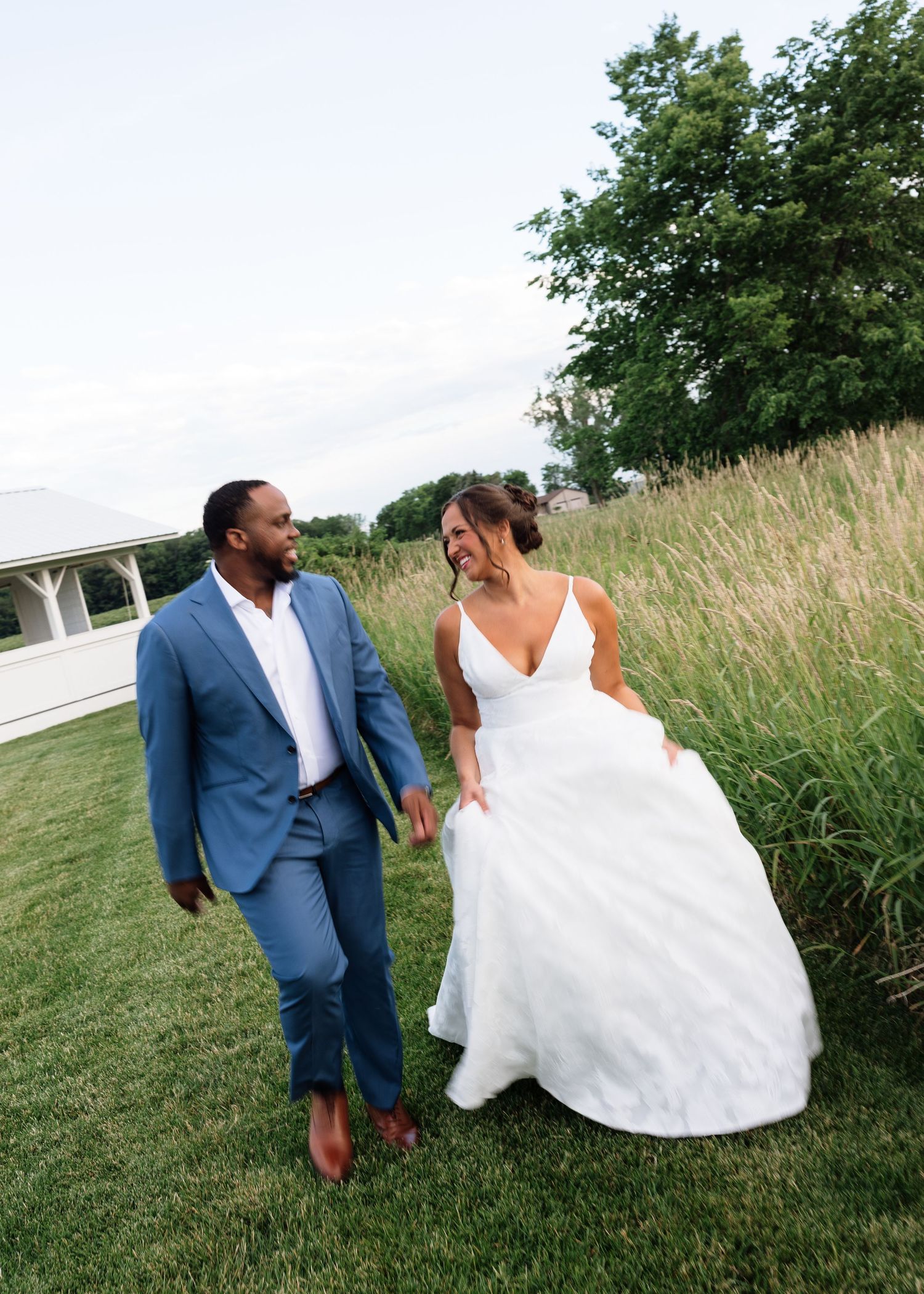 A bride in a white gown and groom in a blue suit walk hand in hand through a lush green field.