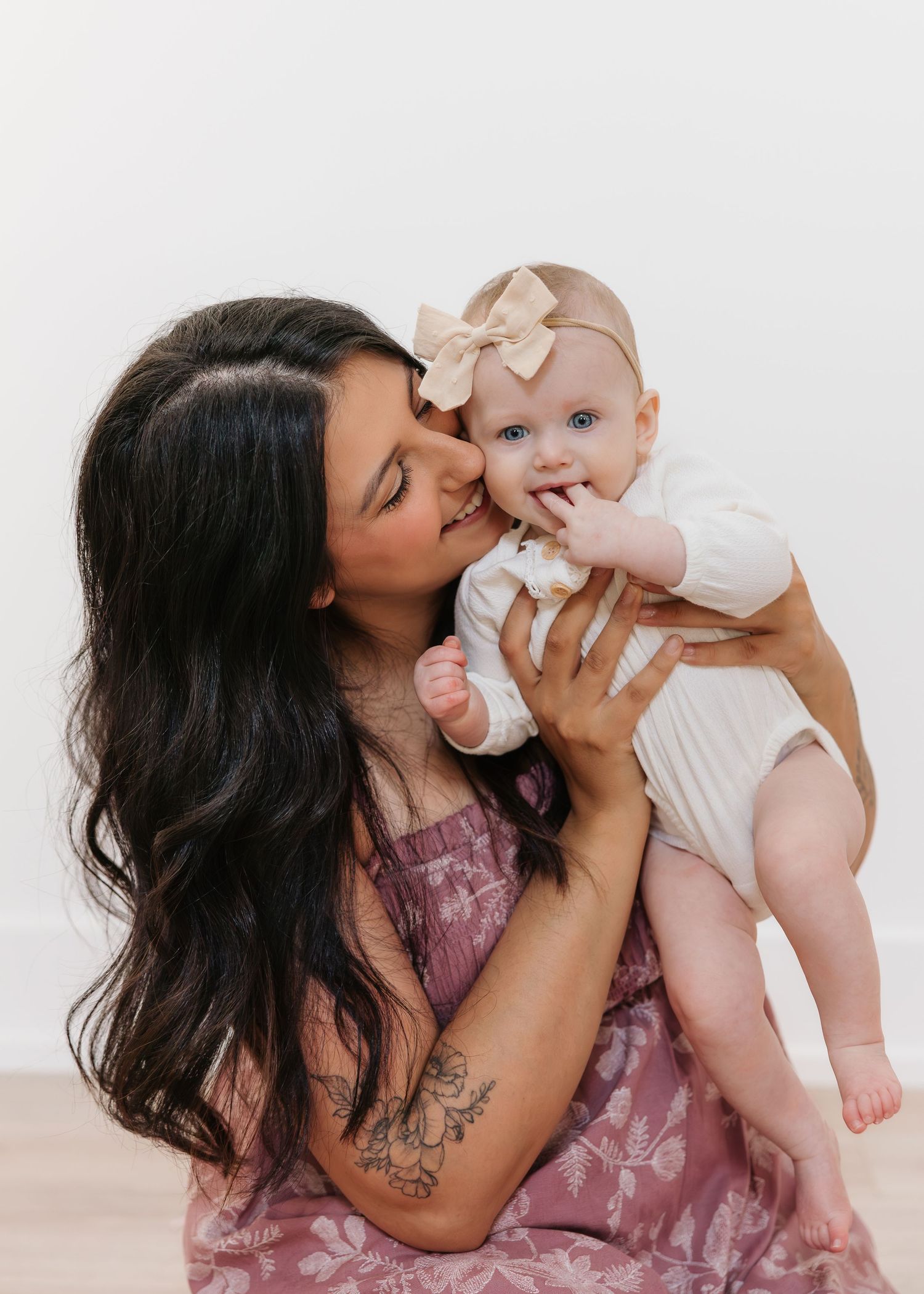 A smiling woman with long dark hair kisses a blue-eyed baby girl wearing a bow headband and white onesie.
