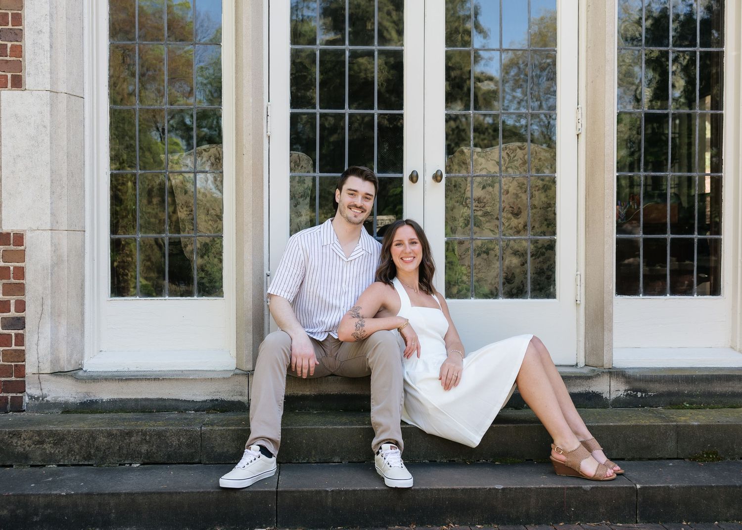 Smiling couple sitting on stone steps in front of white French doors at an outdoor engagement photo session.