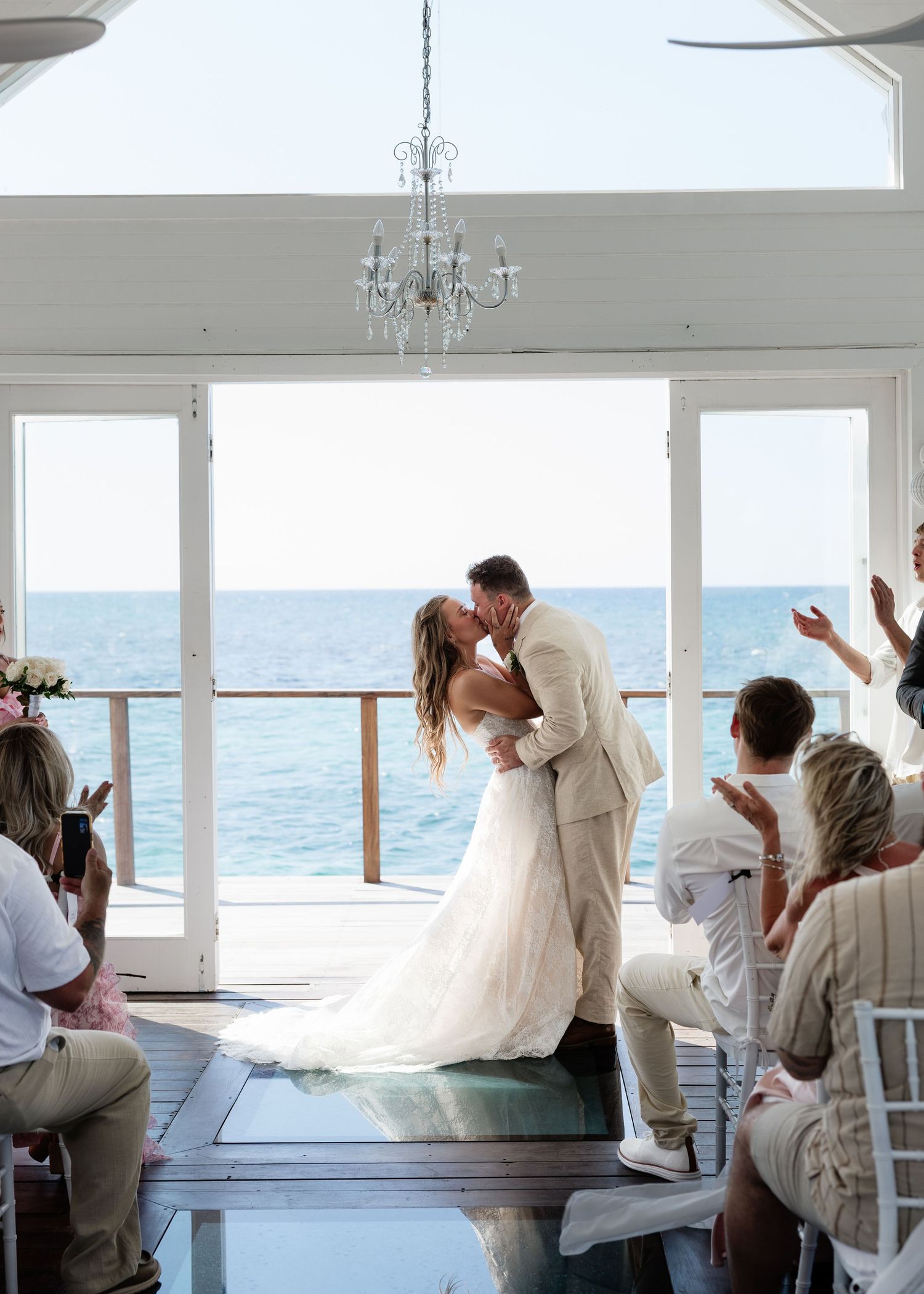A bride and groom share their first kiss at a beachfront wedding ceremony with ocean views and guests applauding.