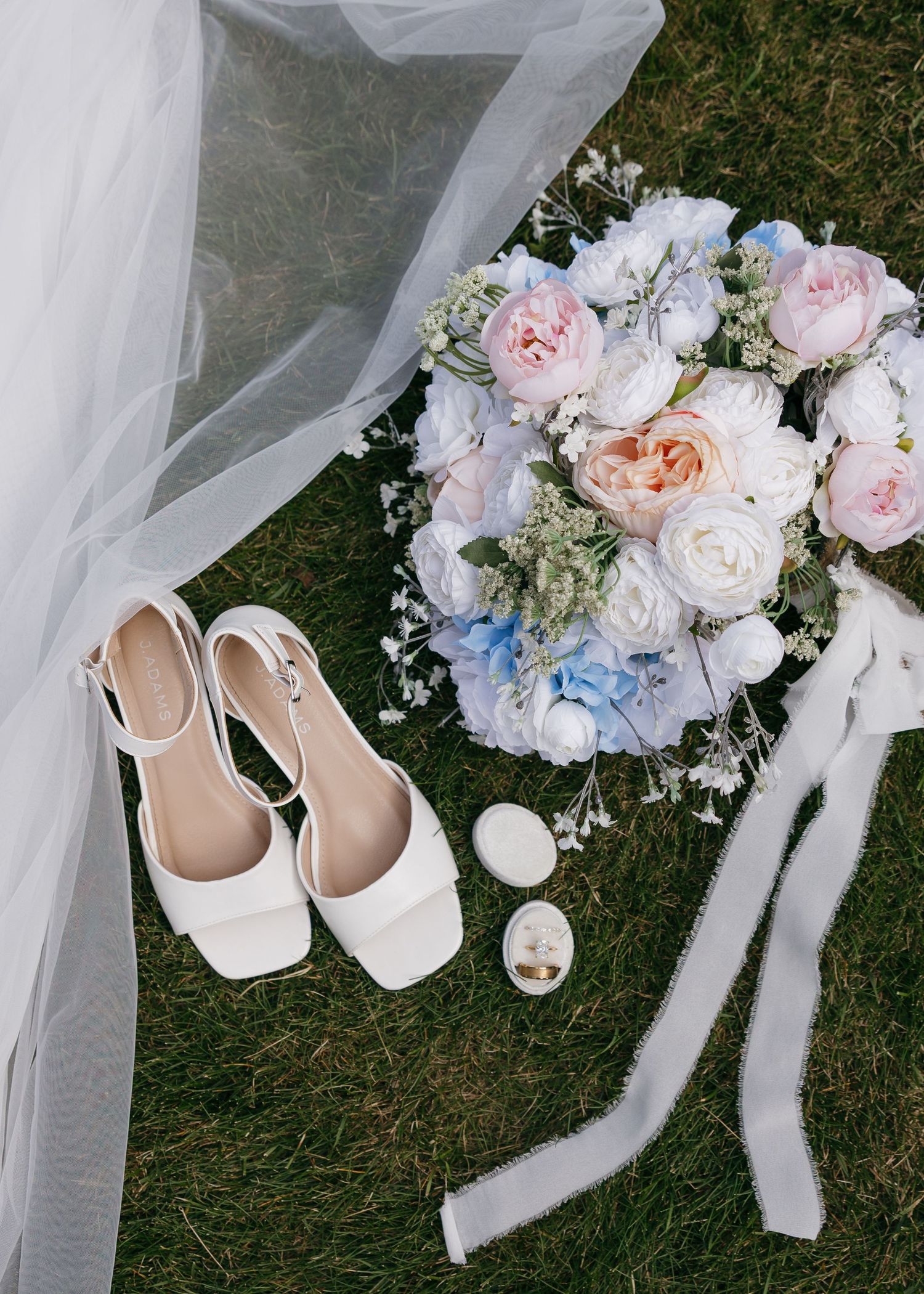 Wedding accessories including white shoes, delicate veil, and a pastel bouquet with roses on grass.