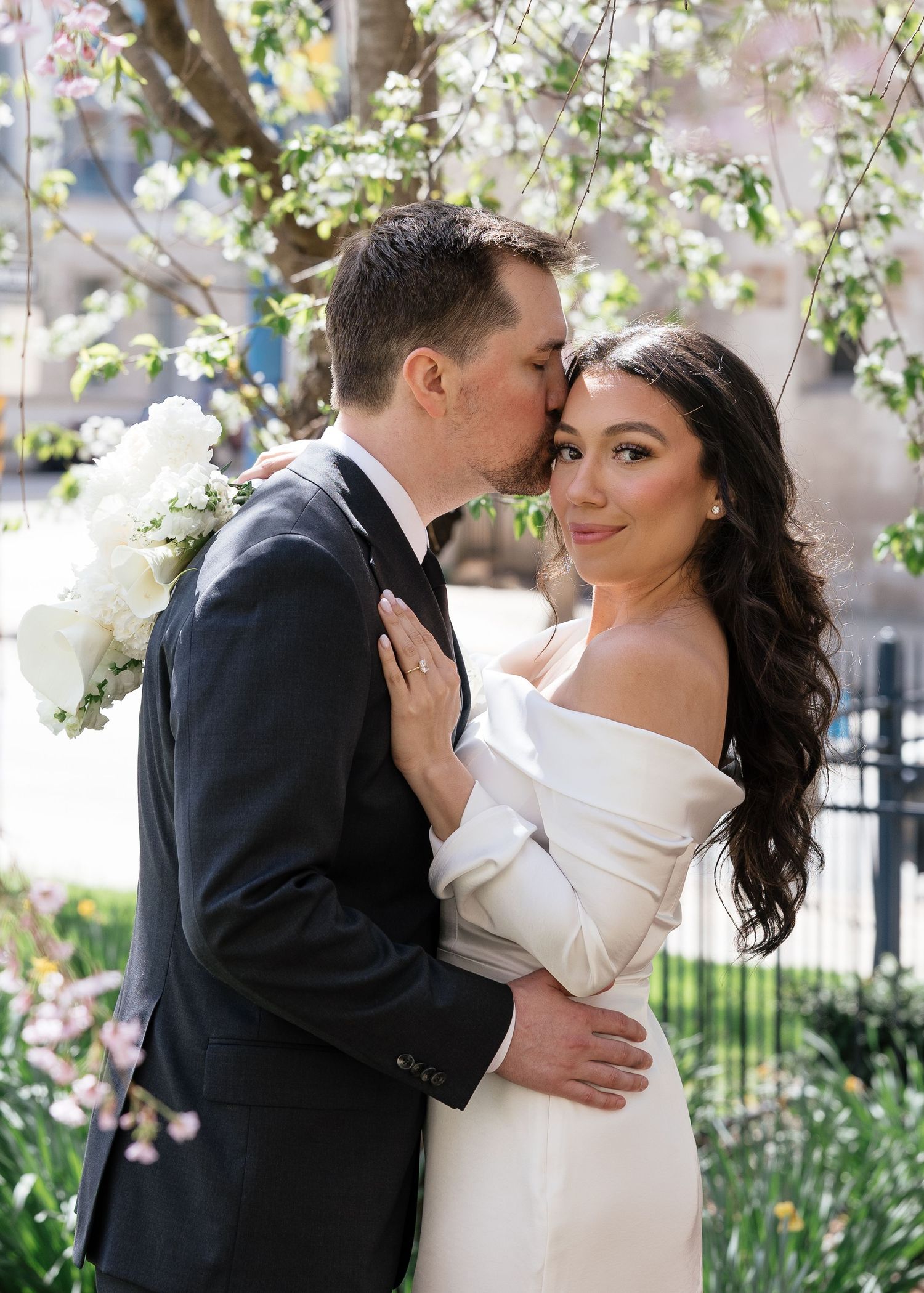 A groom in a black tuxedo kisses his bride in an off-shoulder white gown under blooming spring trees.