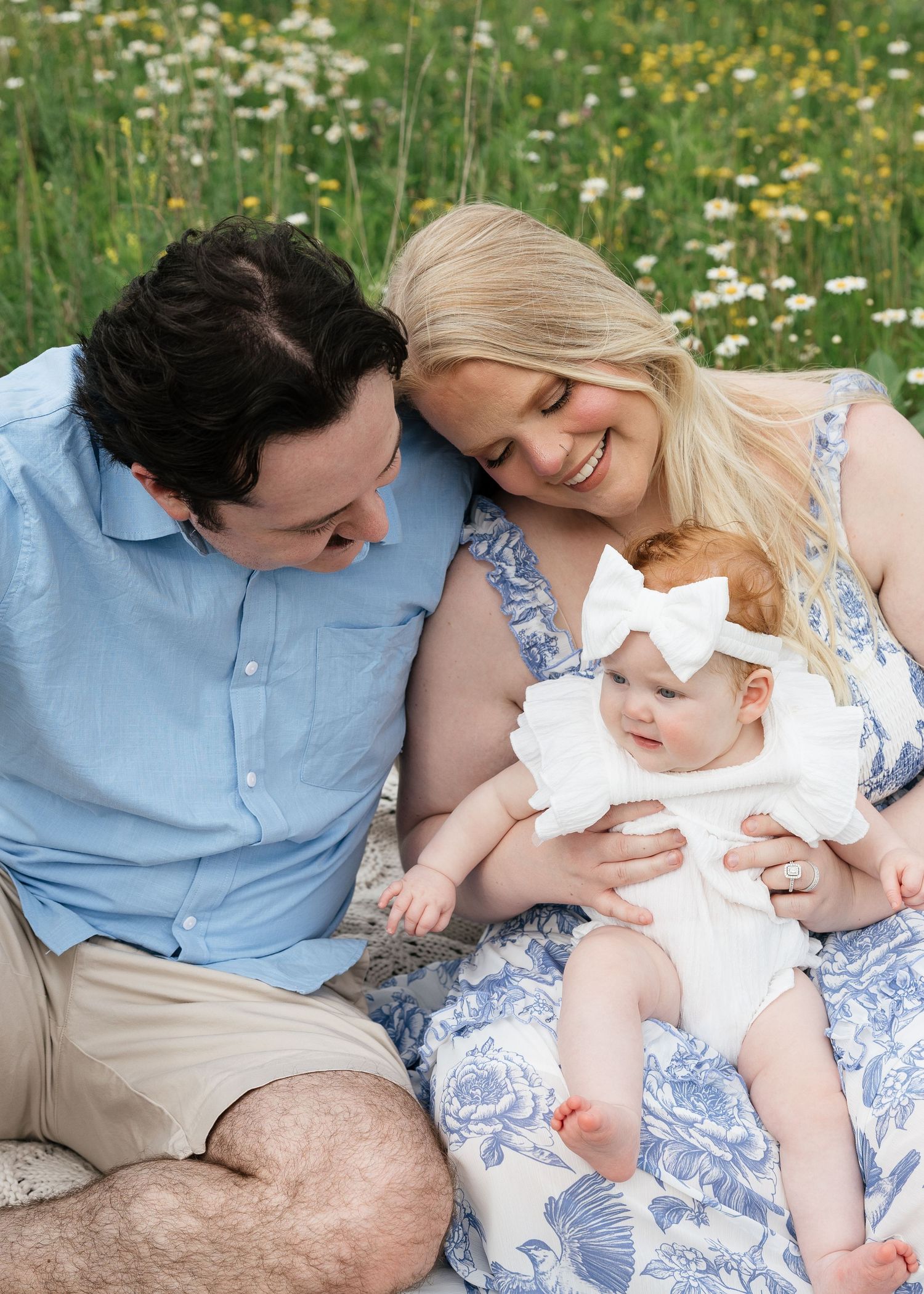 Family photo of parents smiling at baby girl in white dress sitting in sunny wildflower meadow.