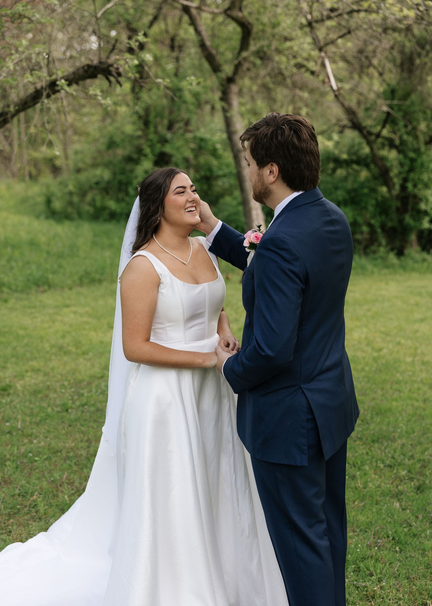 A bride in a white gown and veil smiles as a groom in a navy suit gently touches her face outdoors.