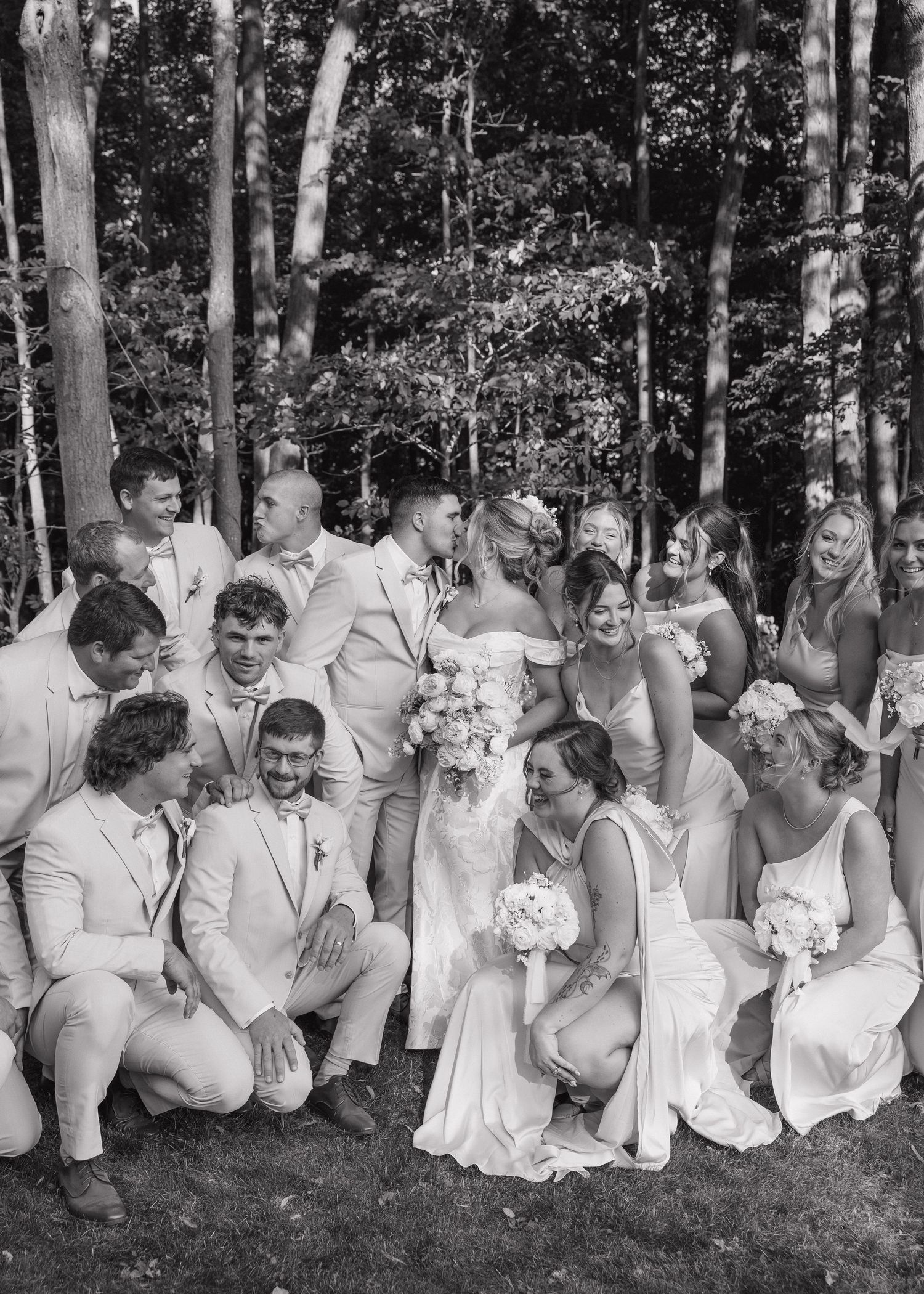 Wedding party in white attire sitting together on grass in front of wooded area.