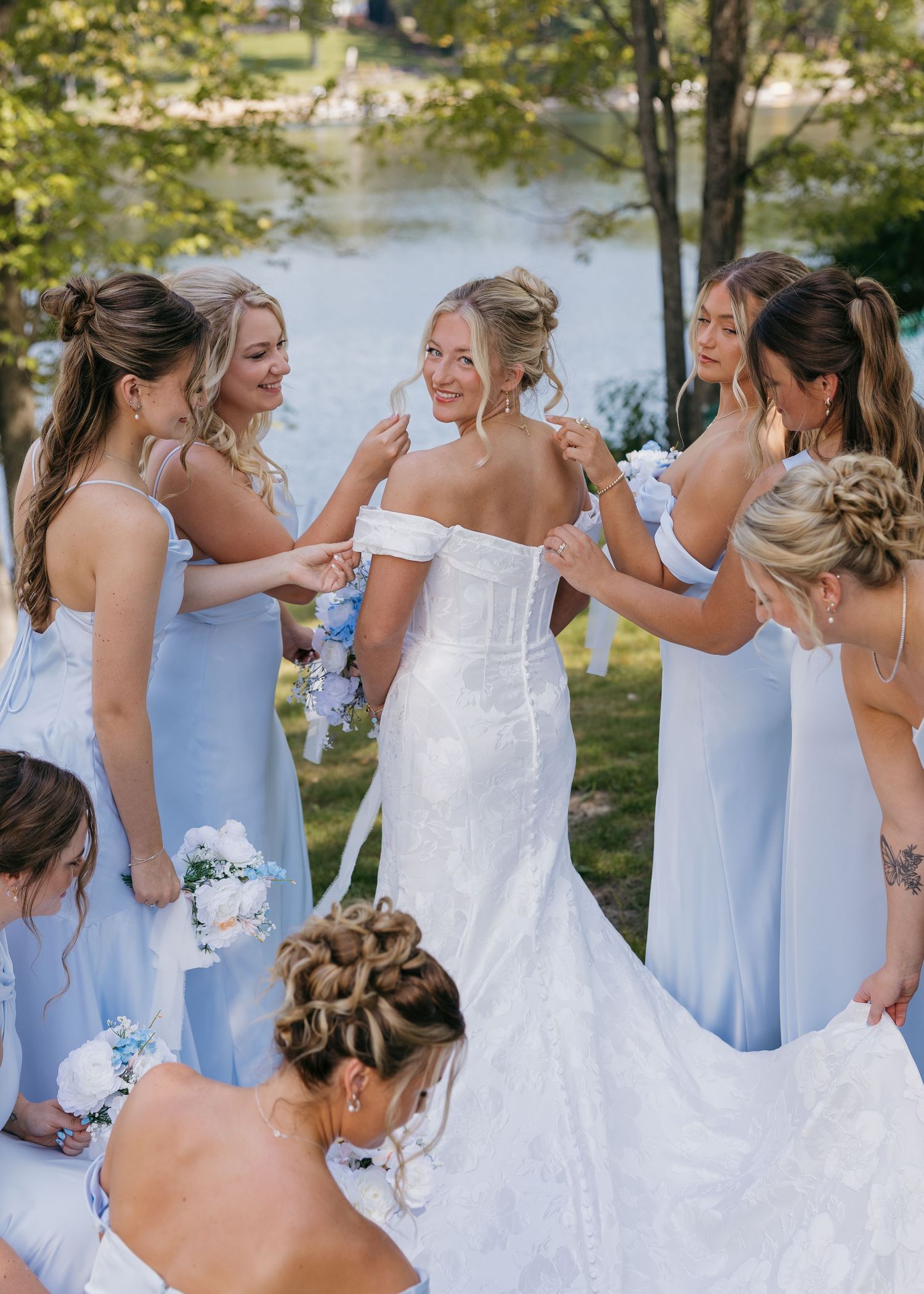 Wedding party in pale blue dresses helping with final dress adjustments by lakeside.