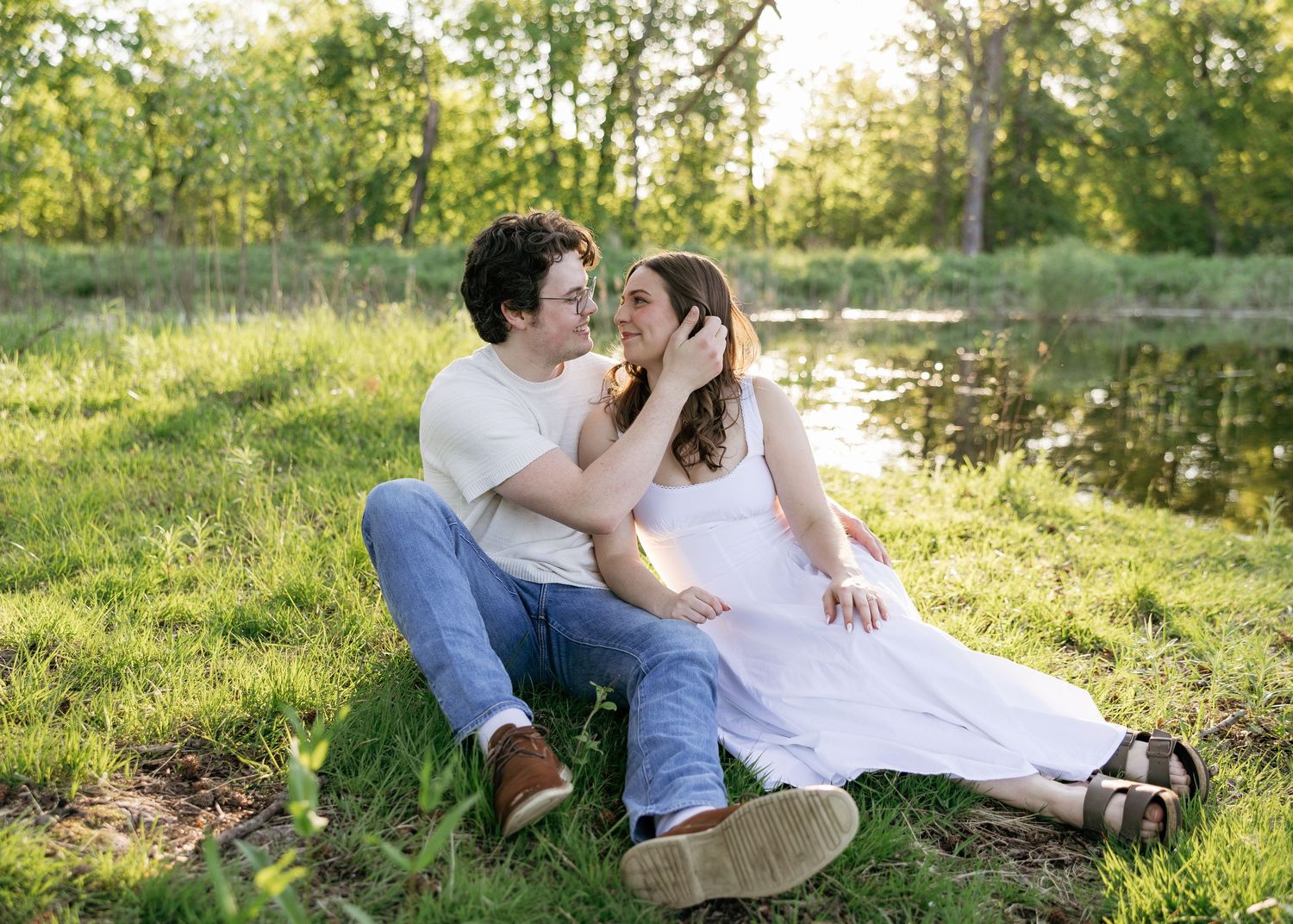 Couple sitting on green grass by a pond, smiling at each other in golden hour light.