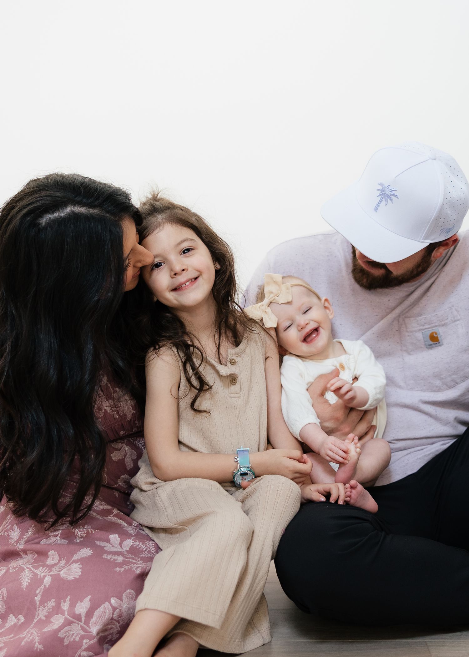 A loving family of four poses together, with mom kissing daughter while dad holds smiling baby, against white background.