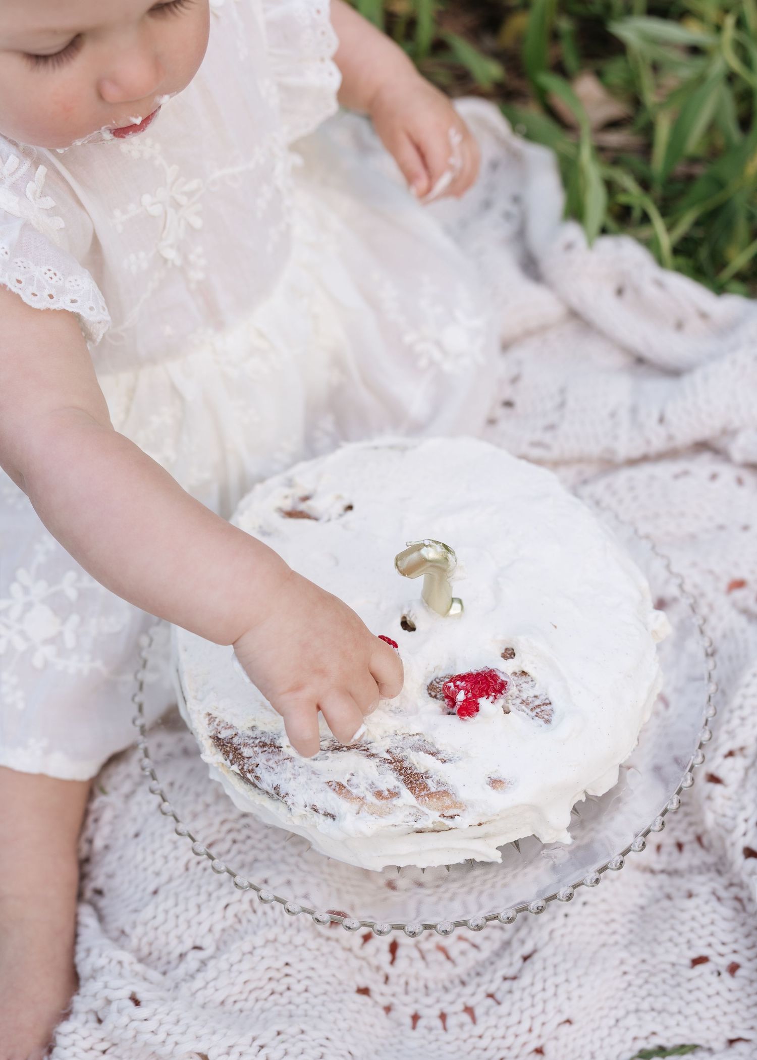 Baby in white dress smashing first birthday cake outdoors on knit blanket.