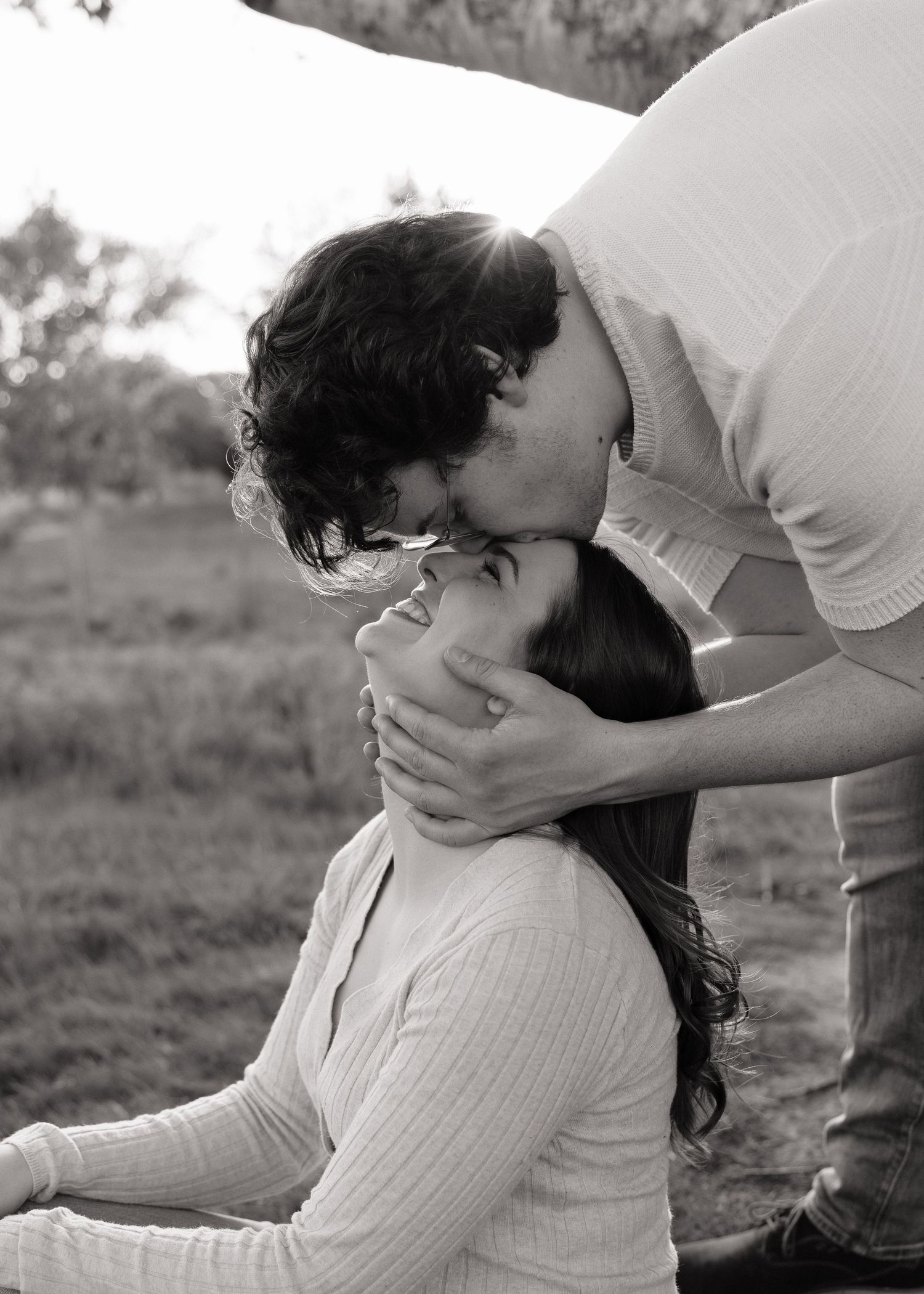 Black and white photo of a man tenderly kissing a woman on the forehead outdoors in a natural setting.