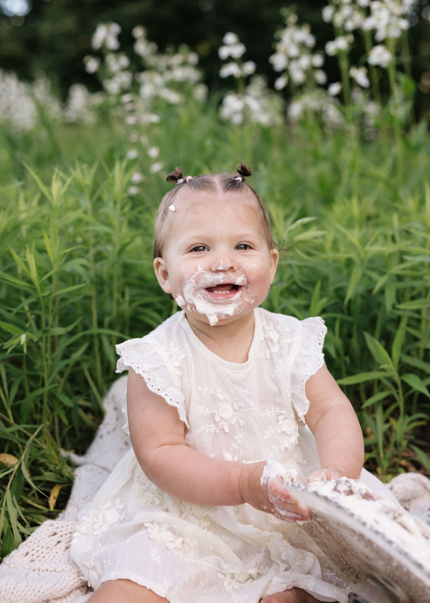 A joyful baby girl in a white floral dress smiles with cake on her face during a first birthday smash cake session.