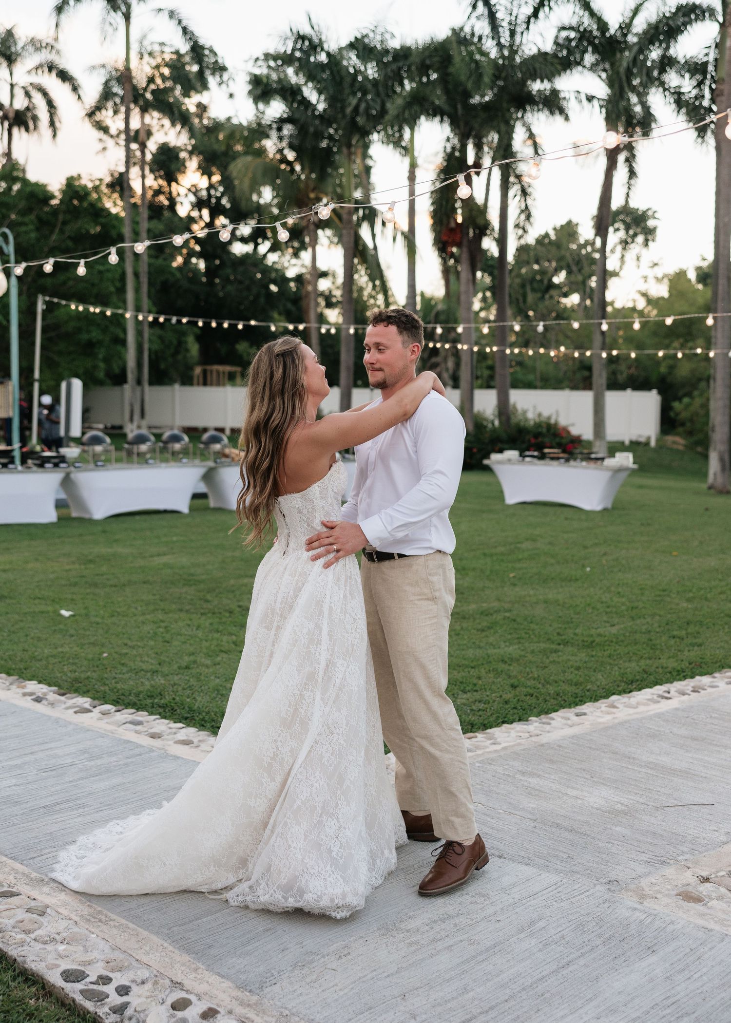 Bride and groom share first dance outdoors at sunset wedding reception with string lights and palm trees.