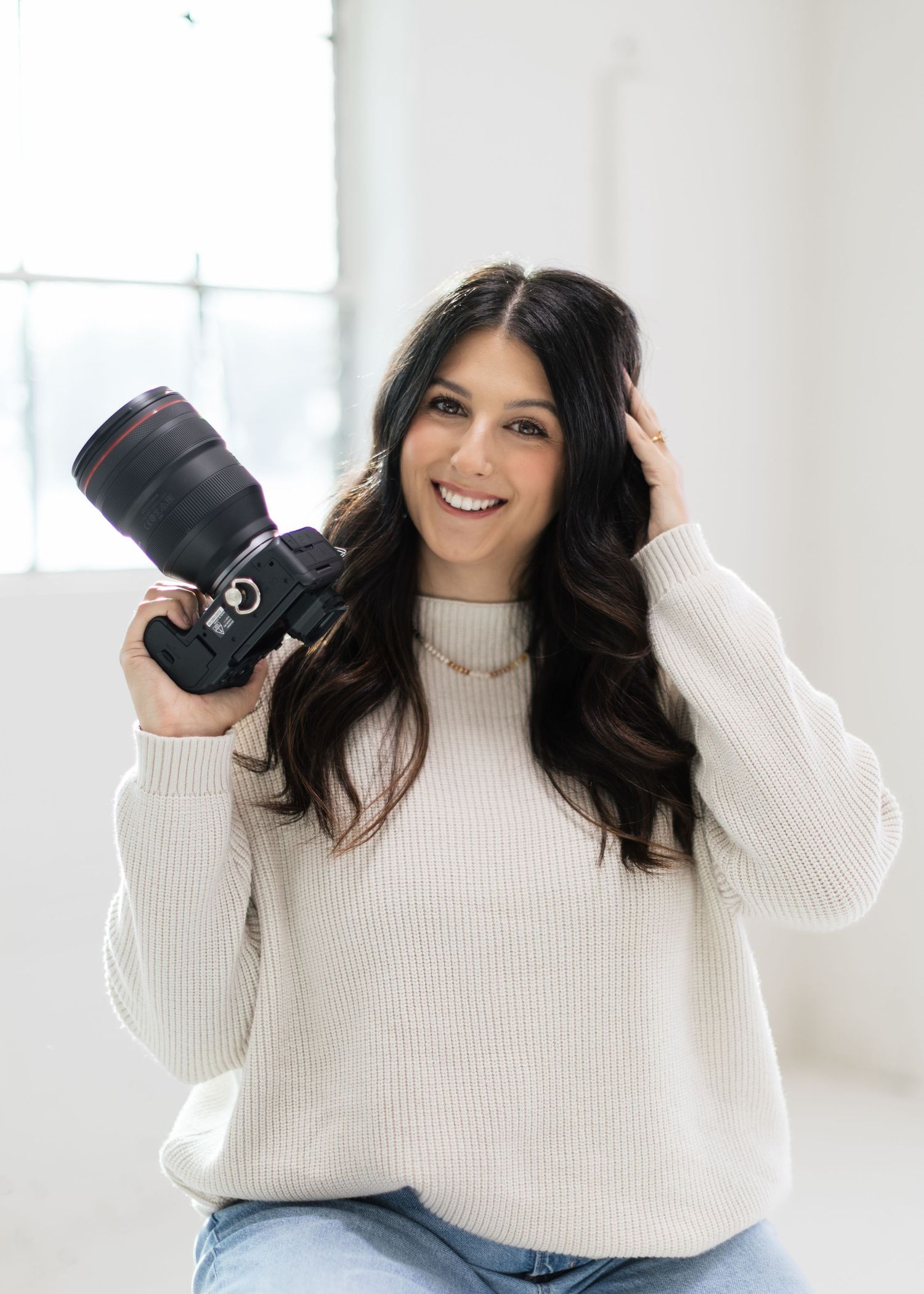 Professional photographer in white sweater posing with camera equipment in bright studio setting.