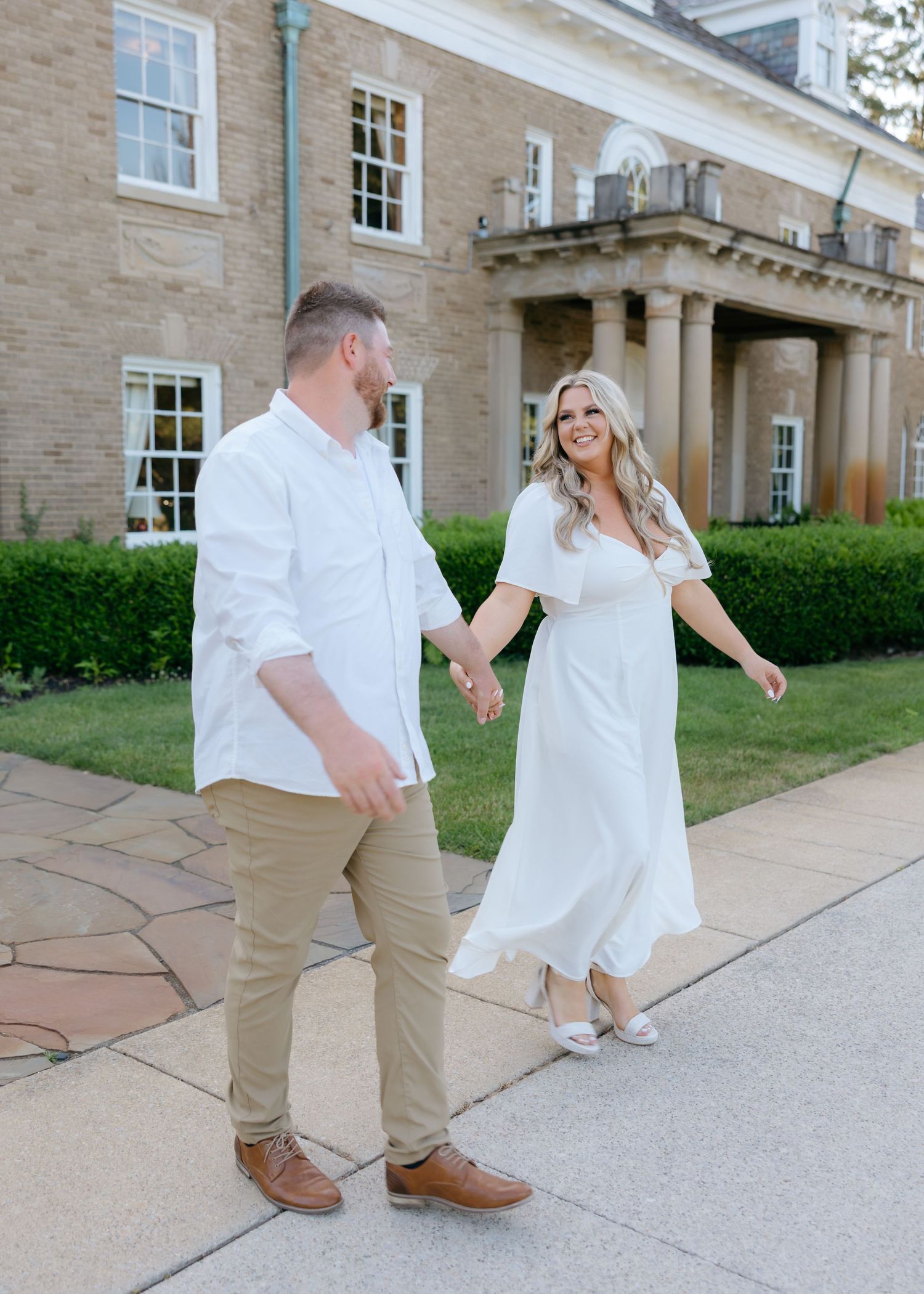 A couple in coordinating white outfits walking together outside a grand stone building.