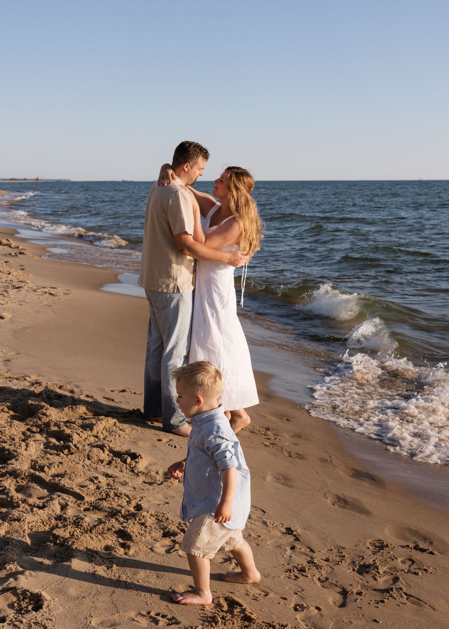 Family of three on sandy beach at sunset, couple embracing near ocean waves while toddler walks barefoot in foreground.