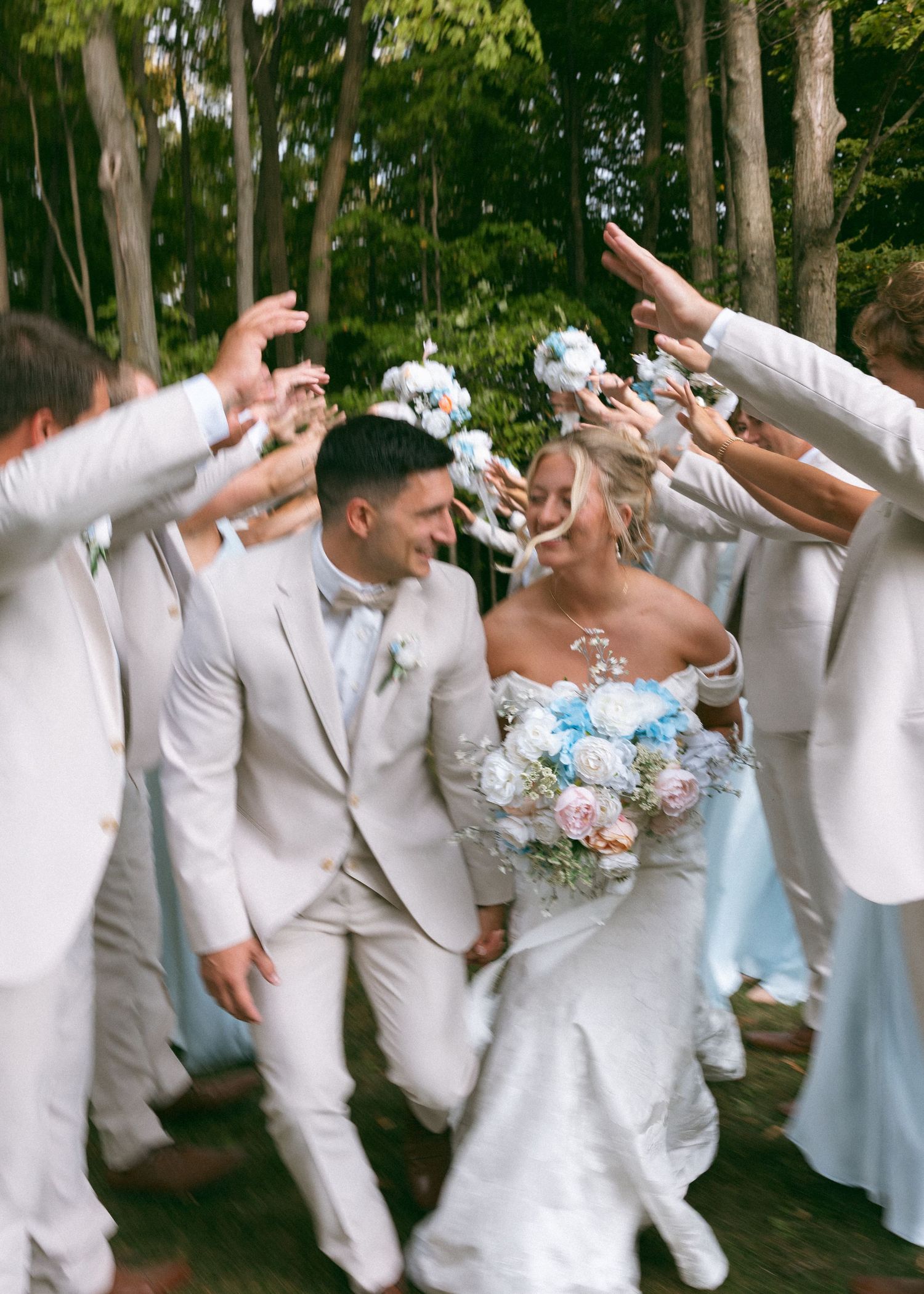 Wedding party celebrates in white attire with joyful poses in a wooded outdoor setting.