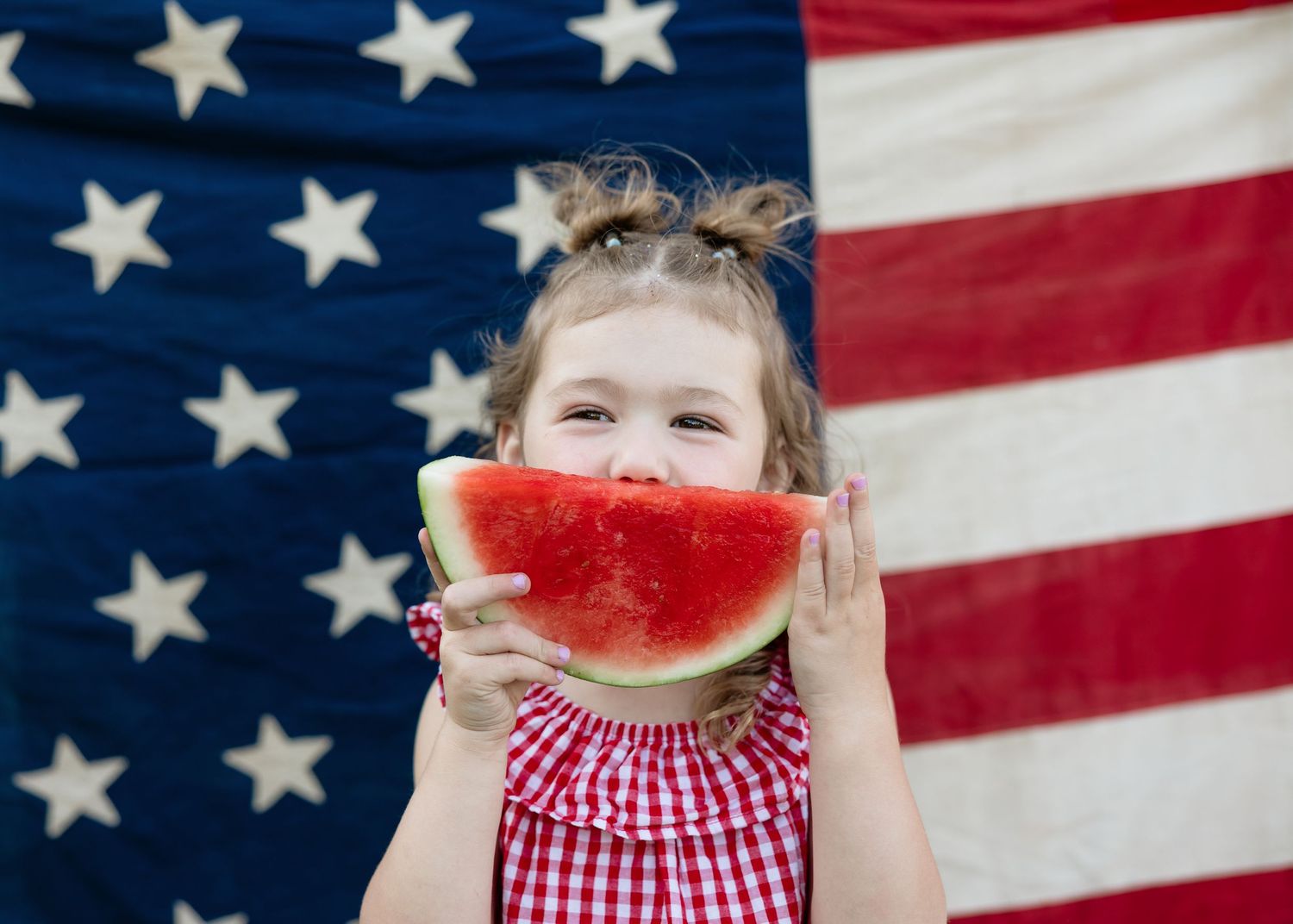 Young girl in red dress eating watermelon slice in front of American flag on Independence Day.