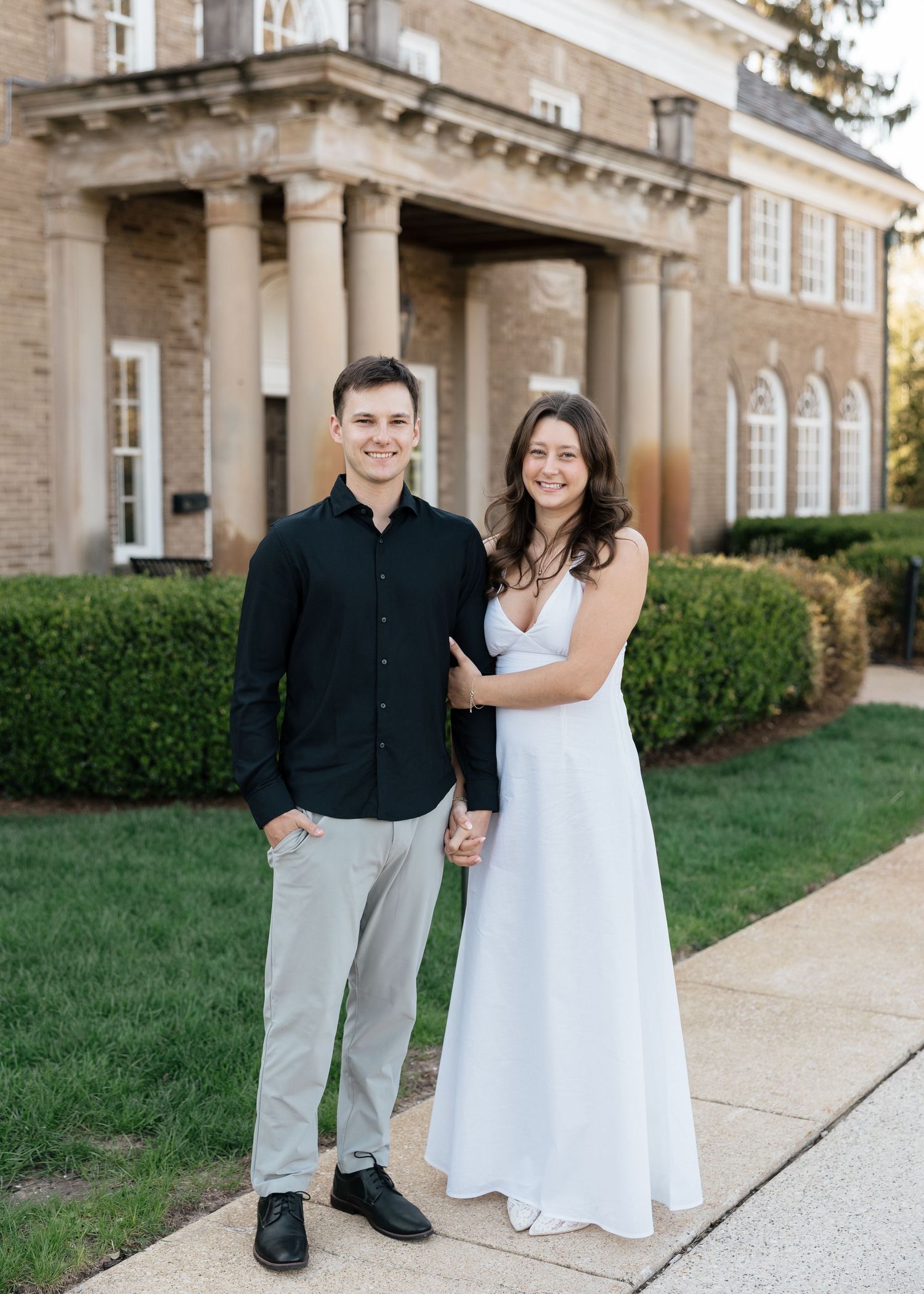 Couple posing in front of a brick building with columns, man in black shirt and woman in white dress.