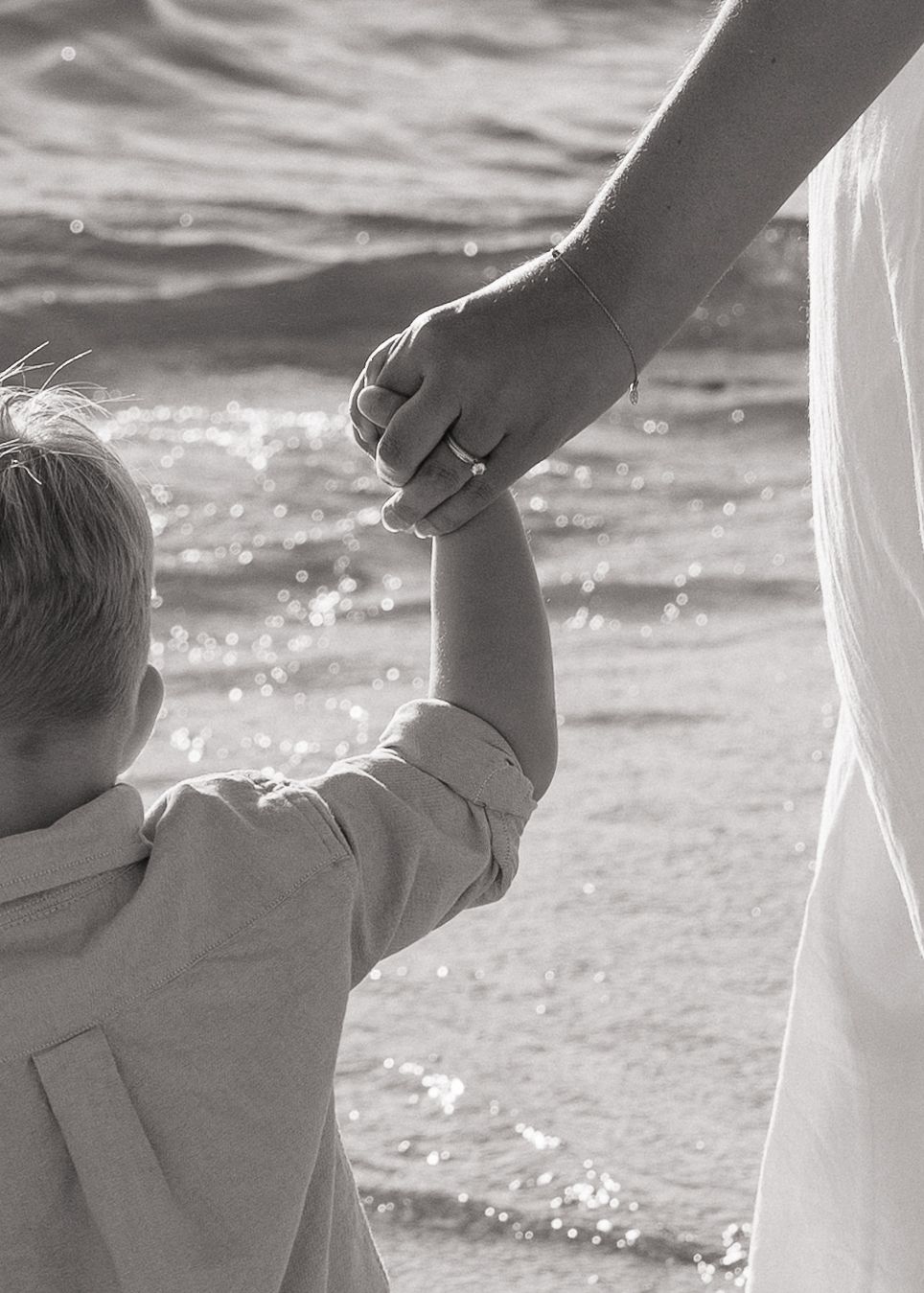 Black and white photo of a young child holding hands with an adult wearing a ring at a sunny beach.