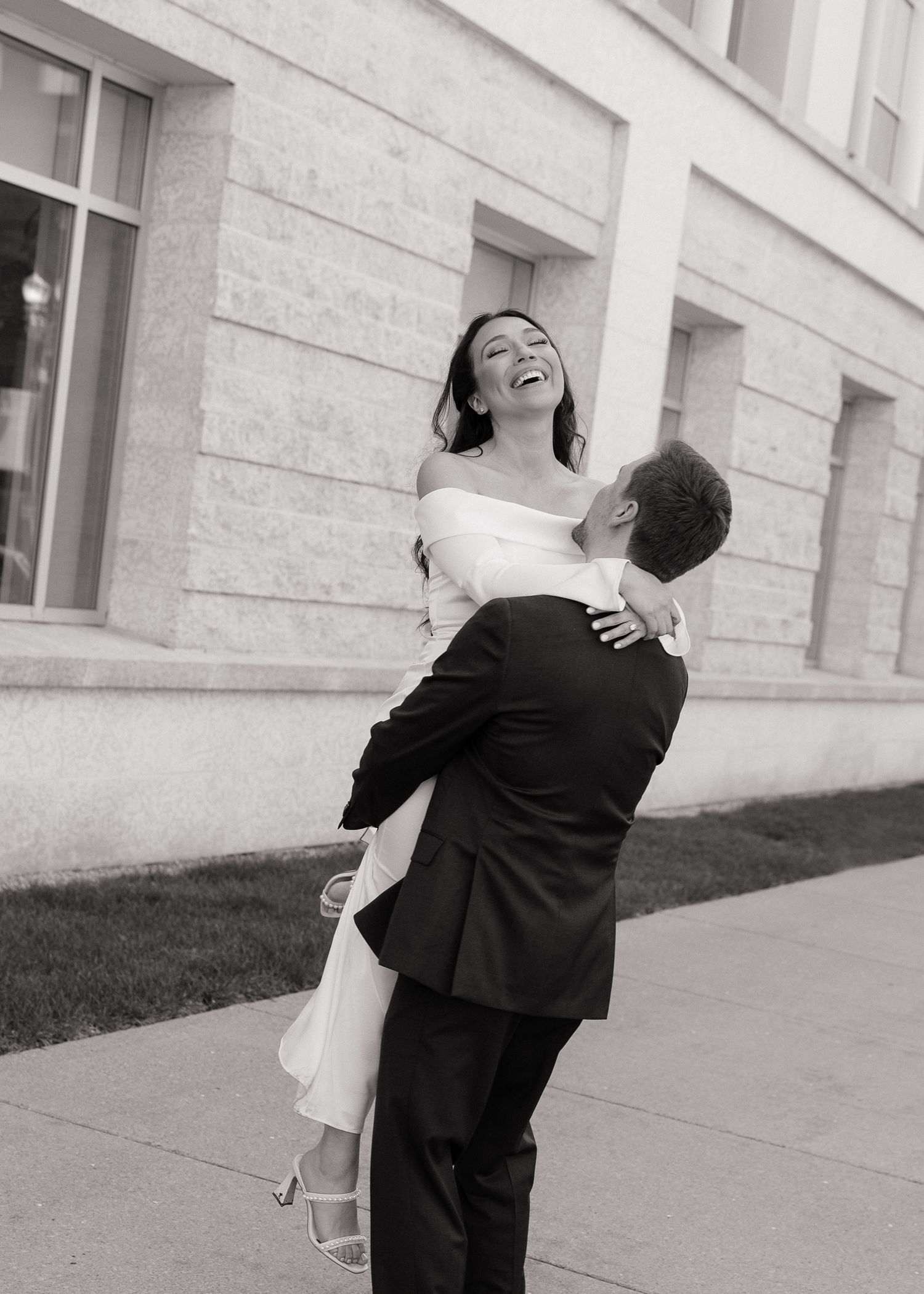 Groom lifting bride in wedding attire, both laughing joyfully outside a stone building, black and white photo.