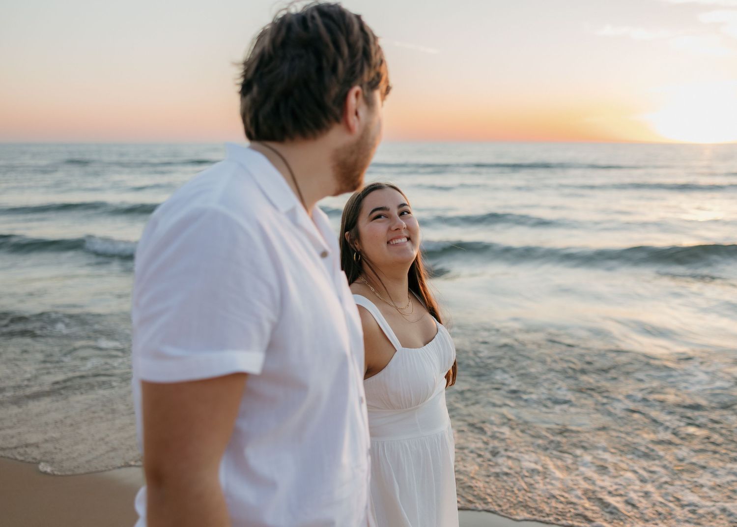 Couple walking along beach at sunset in casual white outfits.